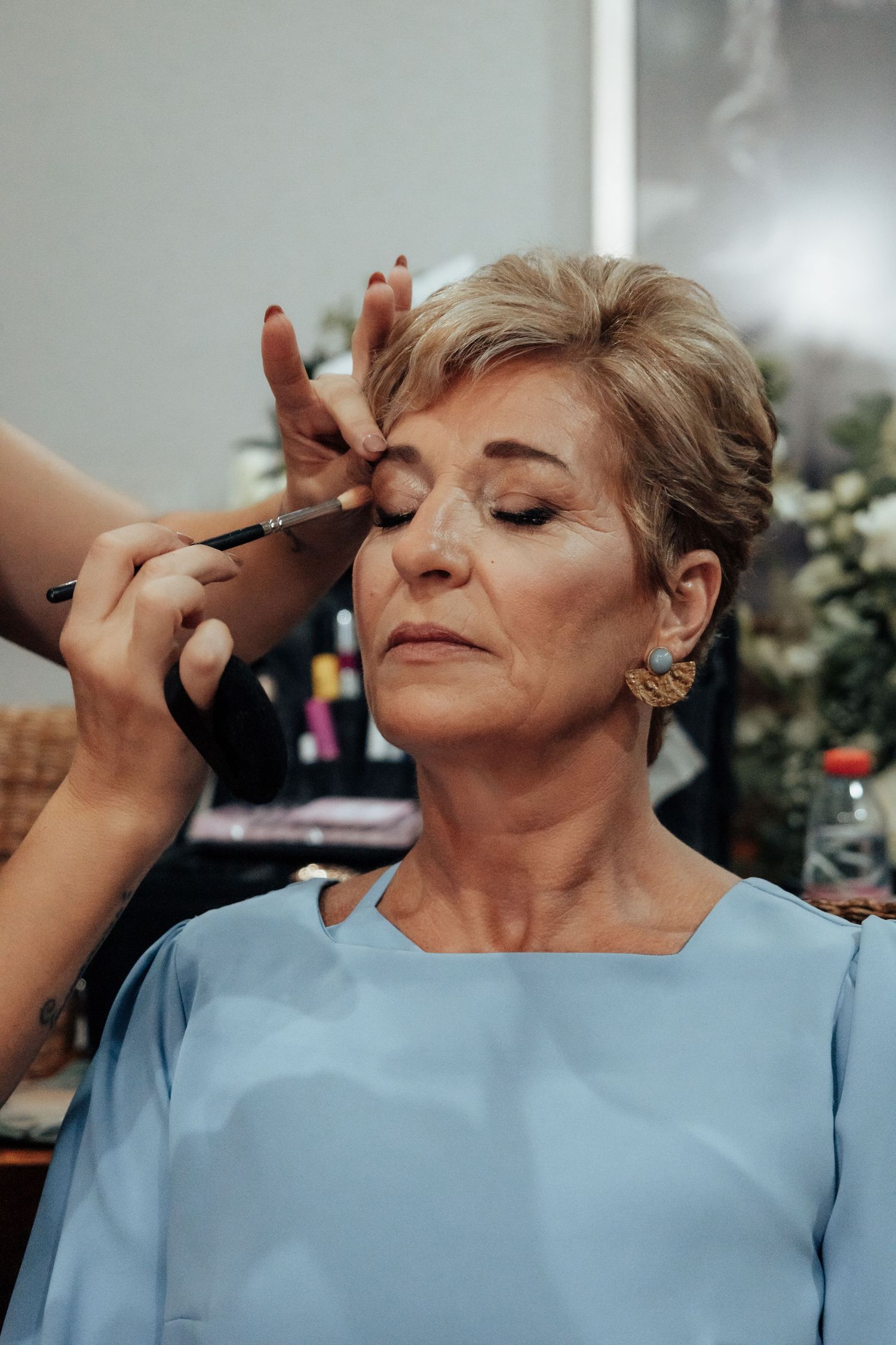 Professional makeup artist applying eyeshadow to an older blonde woman in a blue dress.