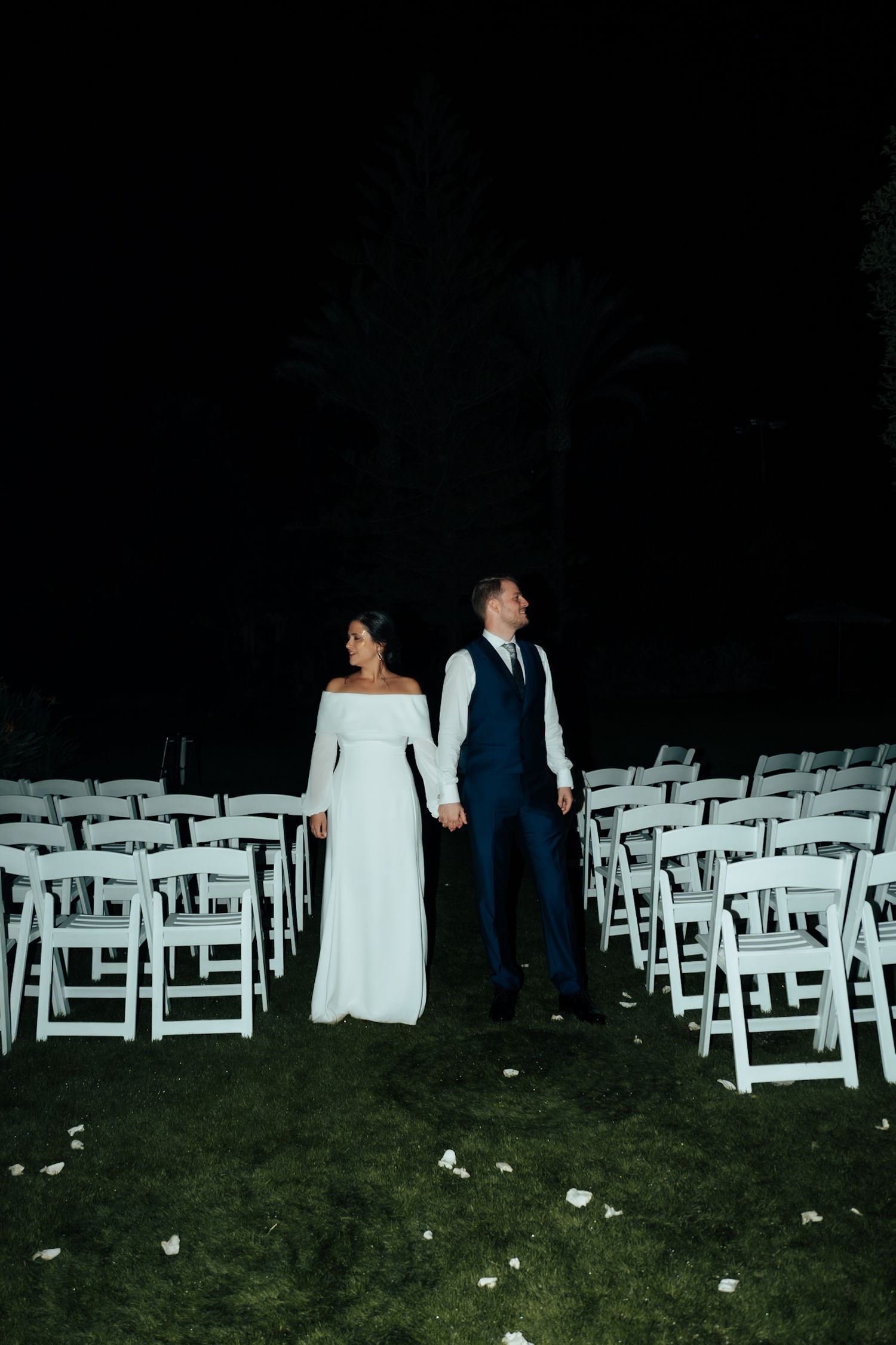 Bride and groom holding hands at night between rows of white folding chairs at outdoor wedding ceremony.