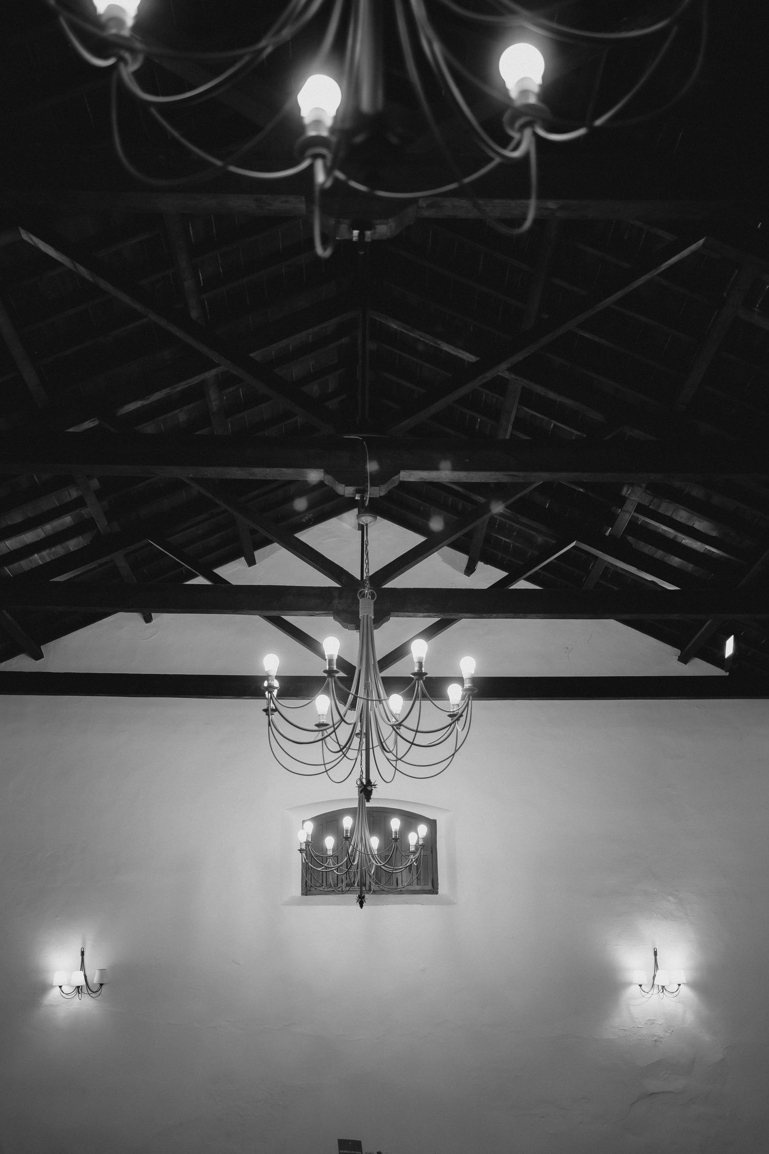 Black and white photo of elegant chandeliers and wall sconces illuminating a rustic wooden beam ceiling.