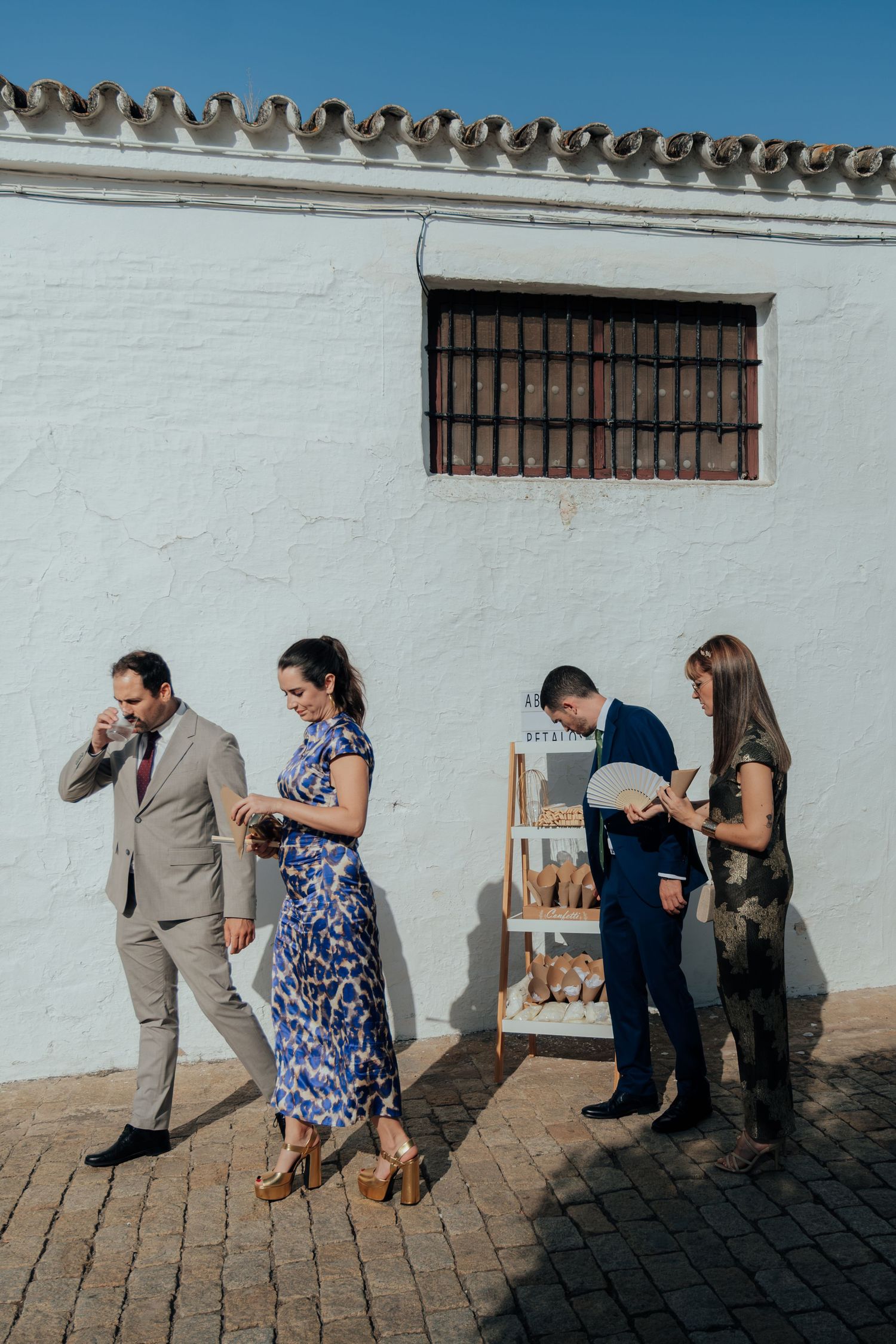Wedding guests socializing outside a whitewashed Spanish venue wall with a wooden display shelf.