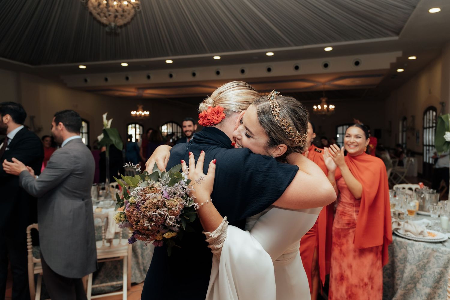 Bride in white gown embraces guest warmly while holding bouquet at elegant wedding reception venue.