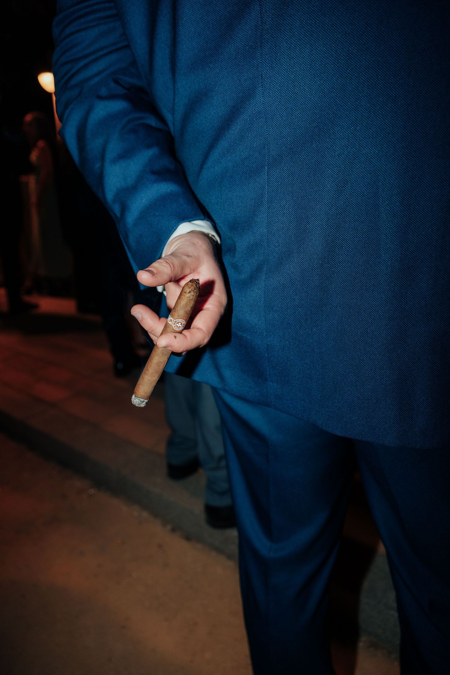 Man in blue suit holding a lit cigar behind his back at a formal evening event.