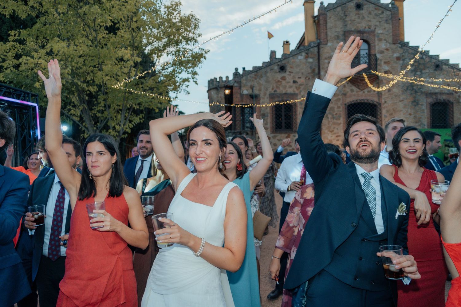 Bride in white gown and groom celebrating with raised hands among wedding guests outside castle.