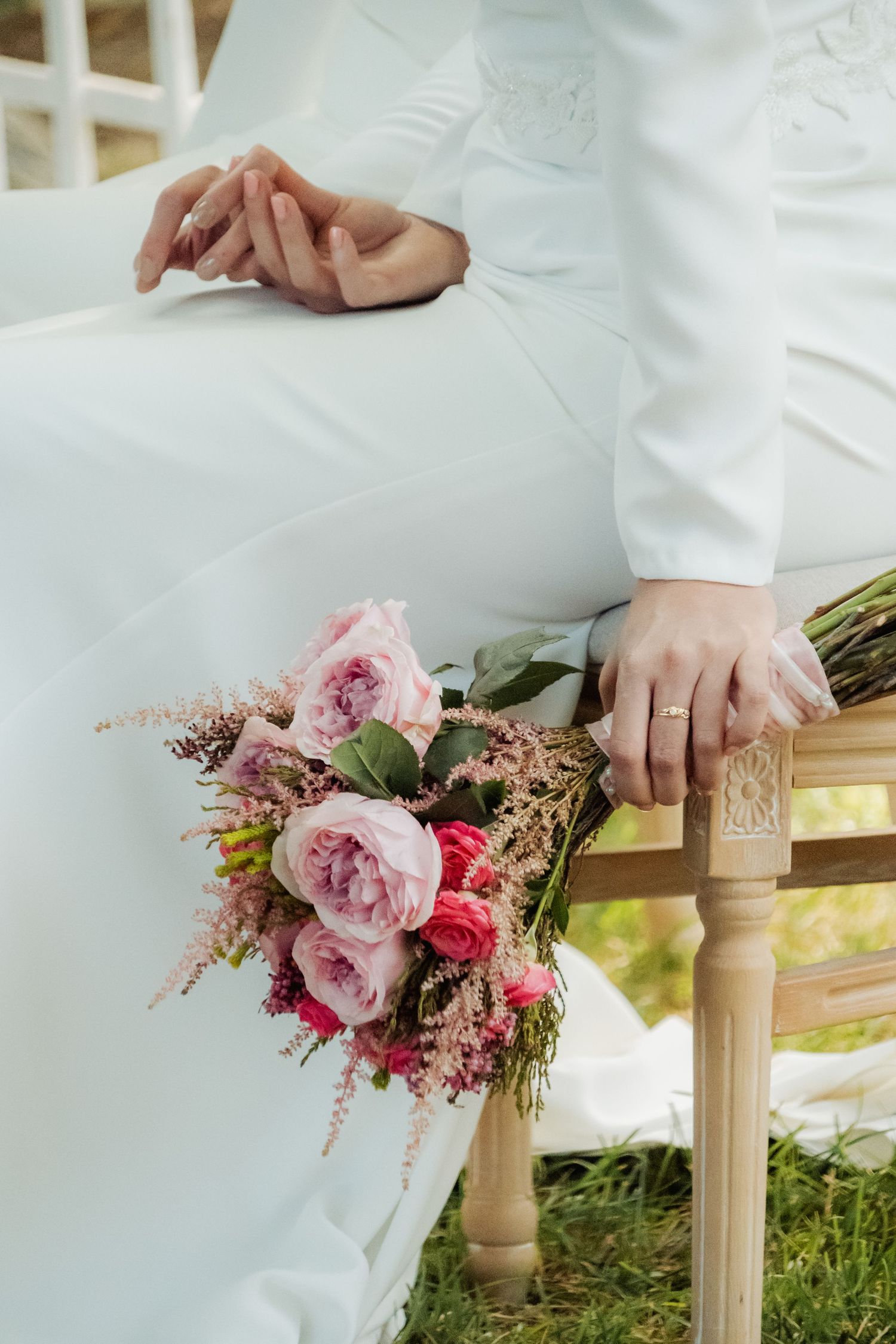Bride in white gown holding a pink peony bouquet on a wooden chair during wedding ceremony.
