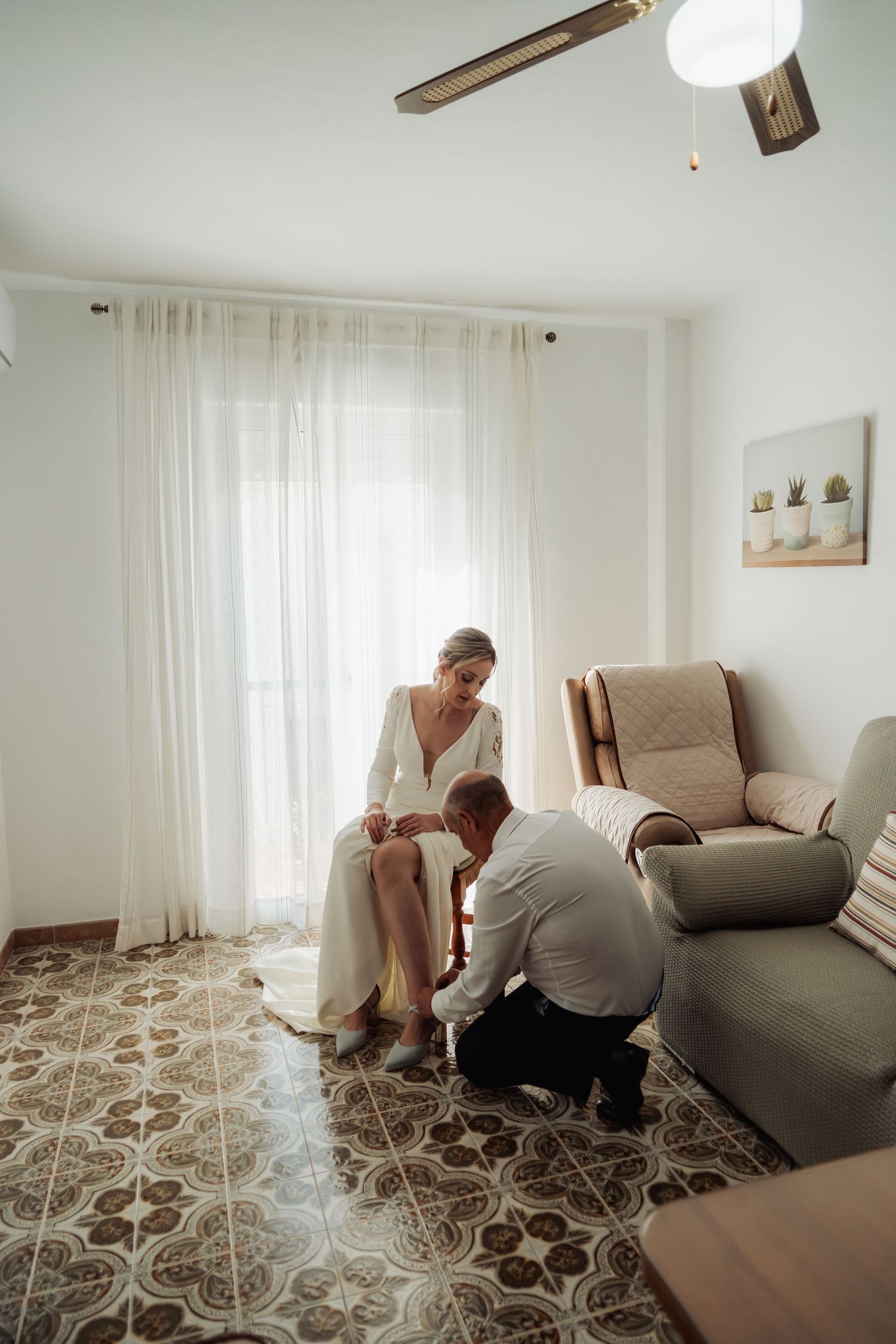 Bride in white gown seated while man helps fasten her shoes in bright tiled room.
