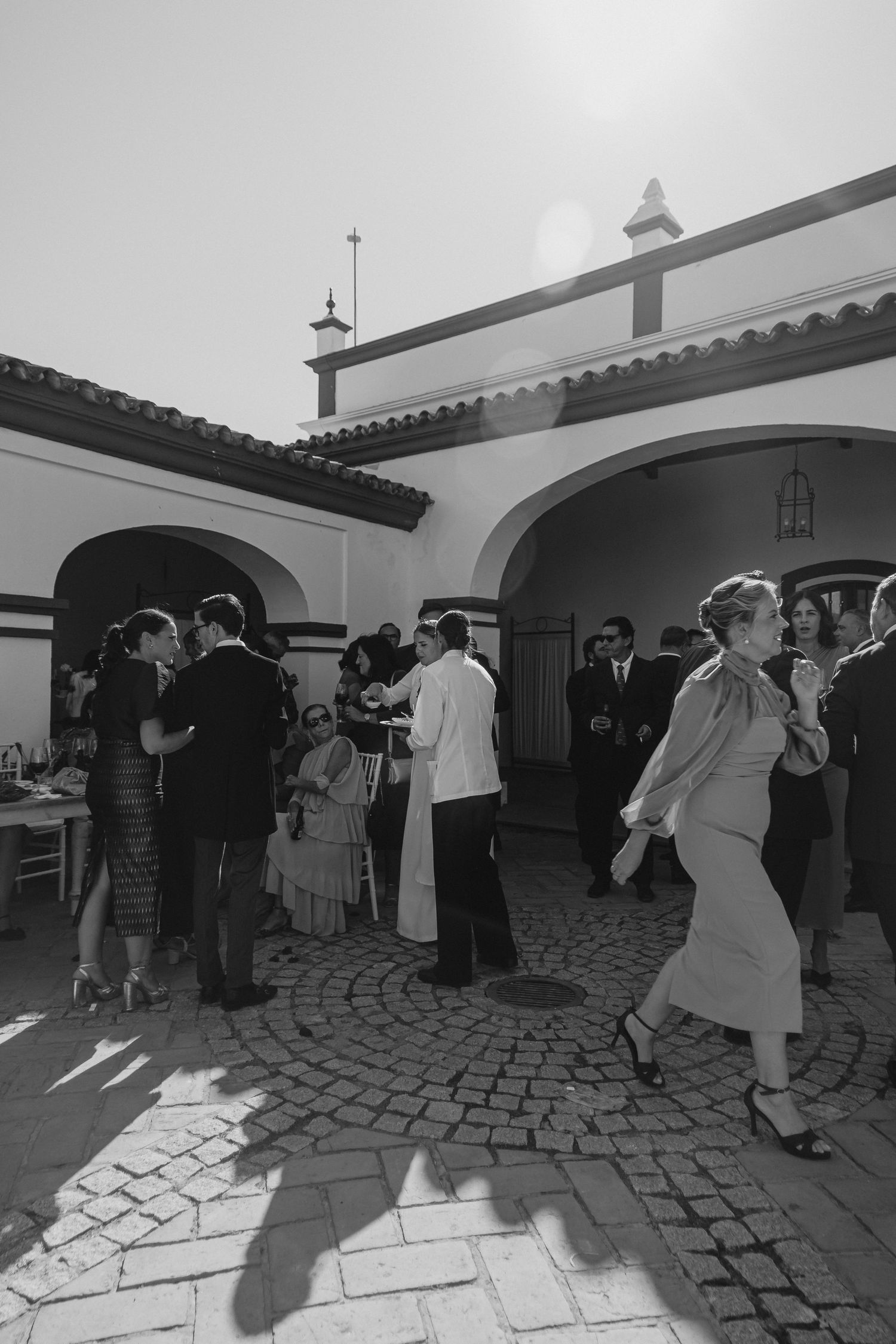 Black and white photo of elegantly dressed guests socializing in a sunlit Spanish courtyard.