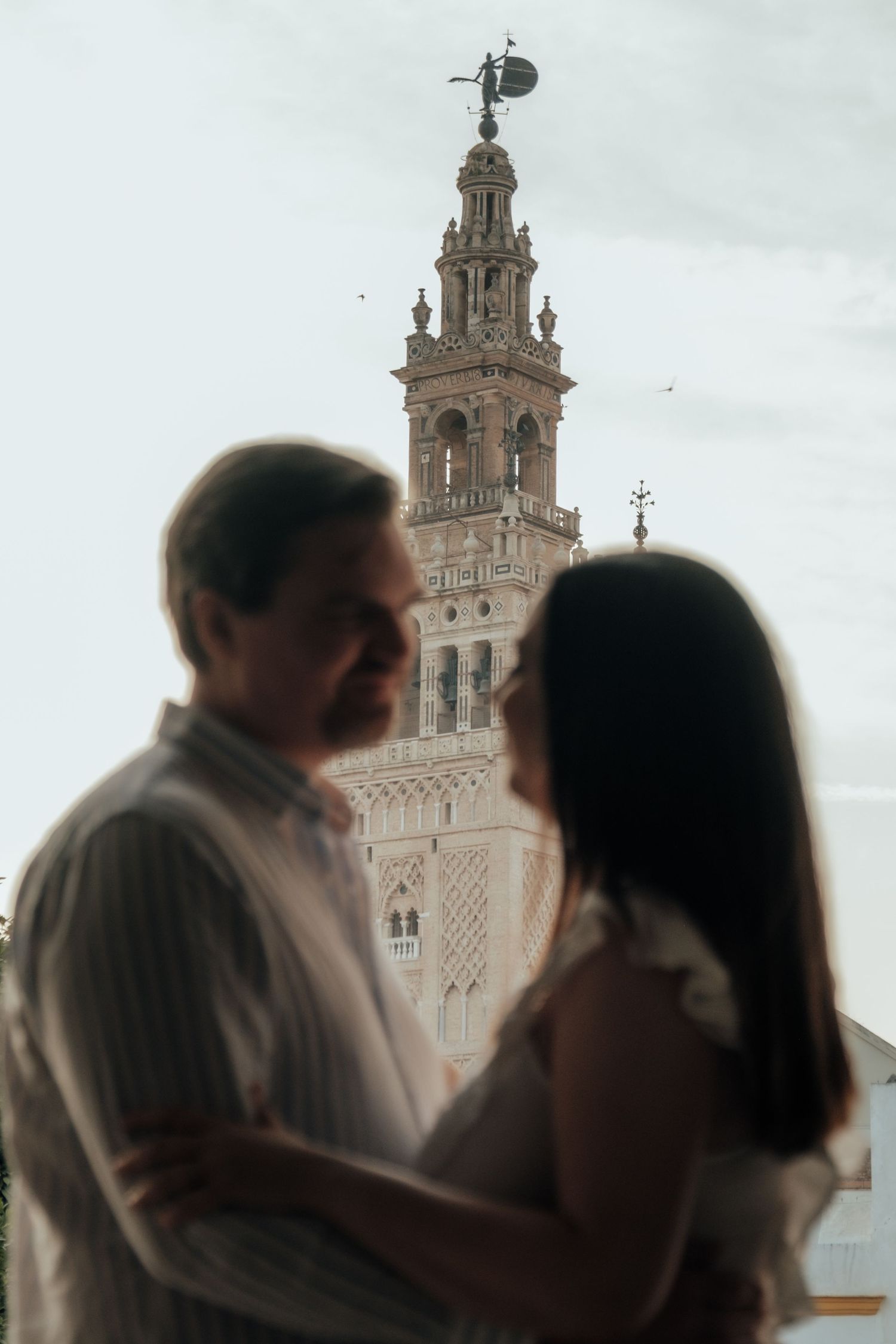 Blurred couple sharing intimate moment with ornate Giralda tower of Seville in background.