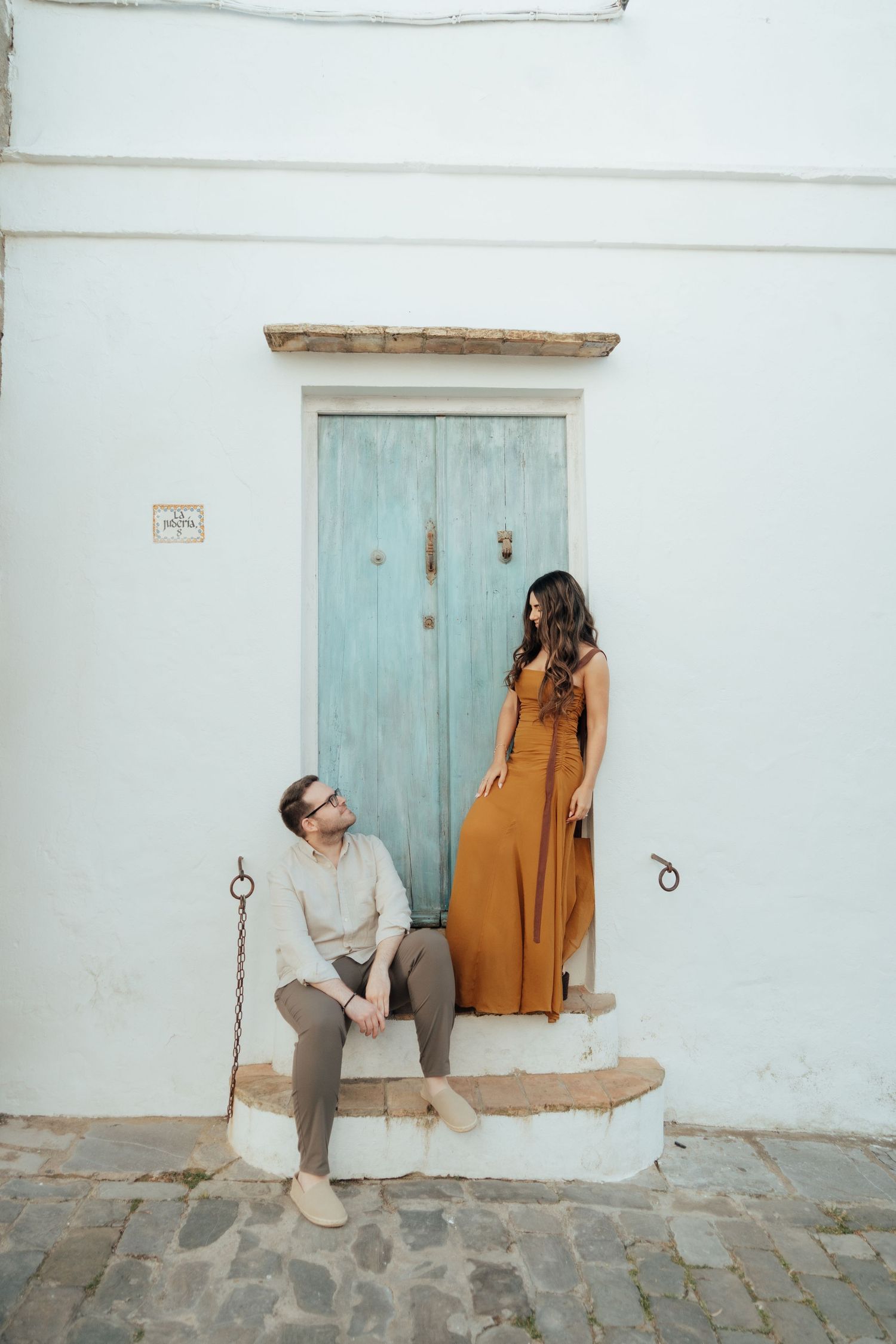 Couple posing by a rustic teal door on white Mediterranean building, woman in mustard dress, man in beige outfit.