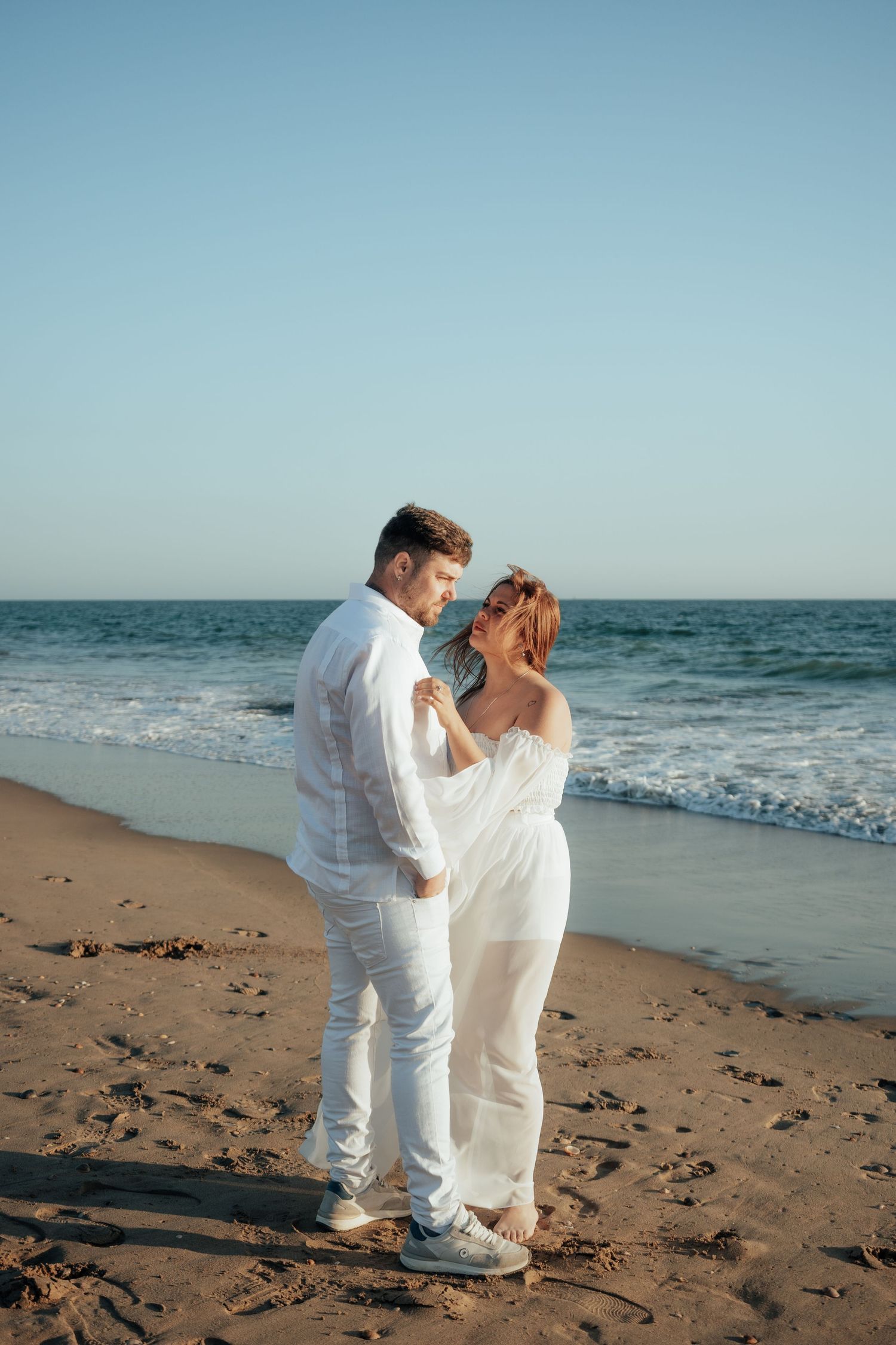 Romantic couple in white outfits embracing on a sandy beach at sunset with ocean waves in the background.