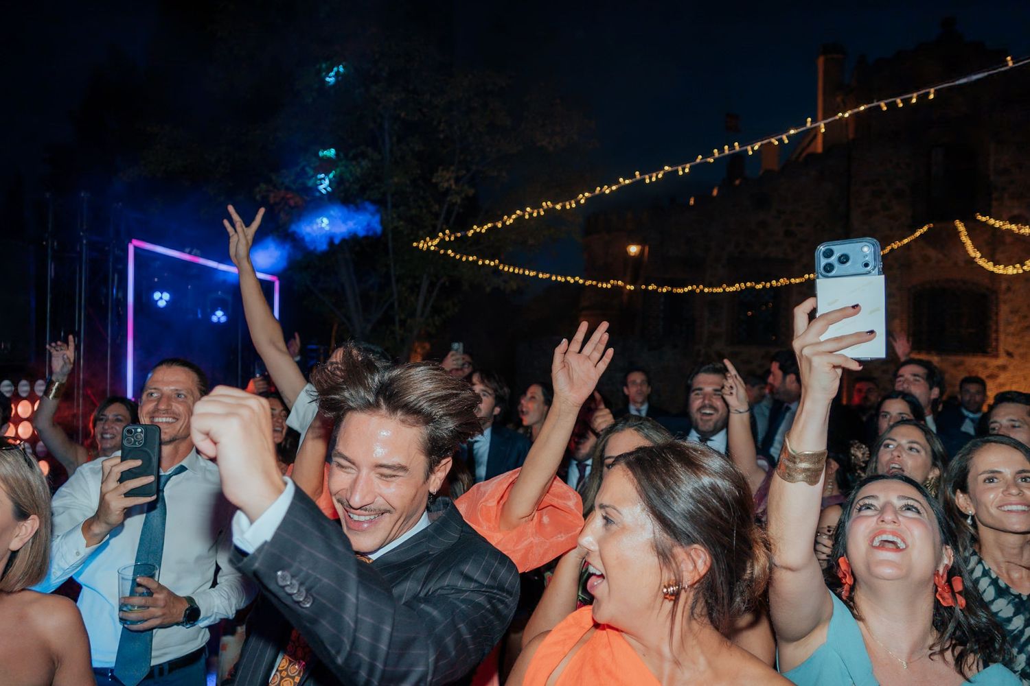 Joyful wedding guests dancing and celebrating with raised arms at lively outdoor night reception.