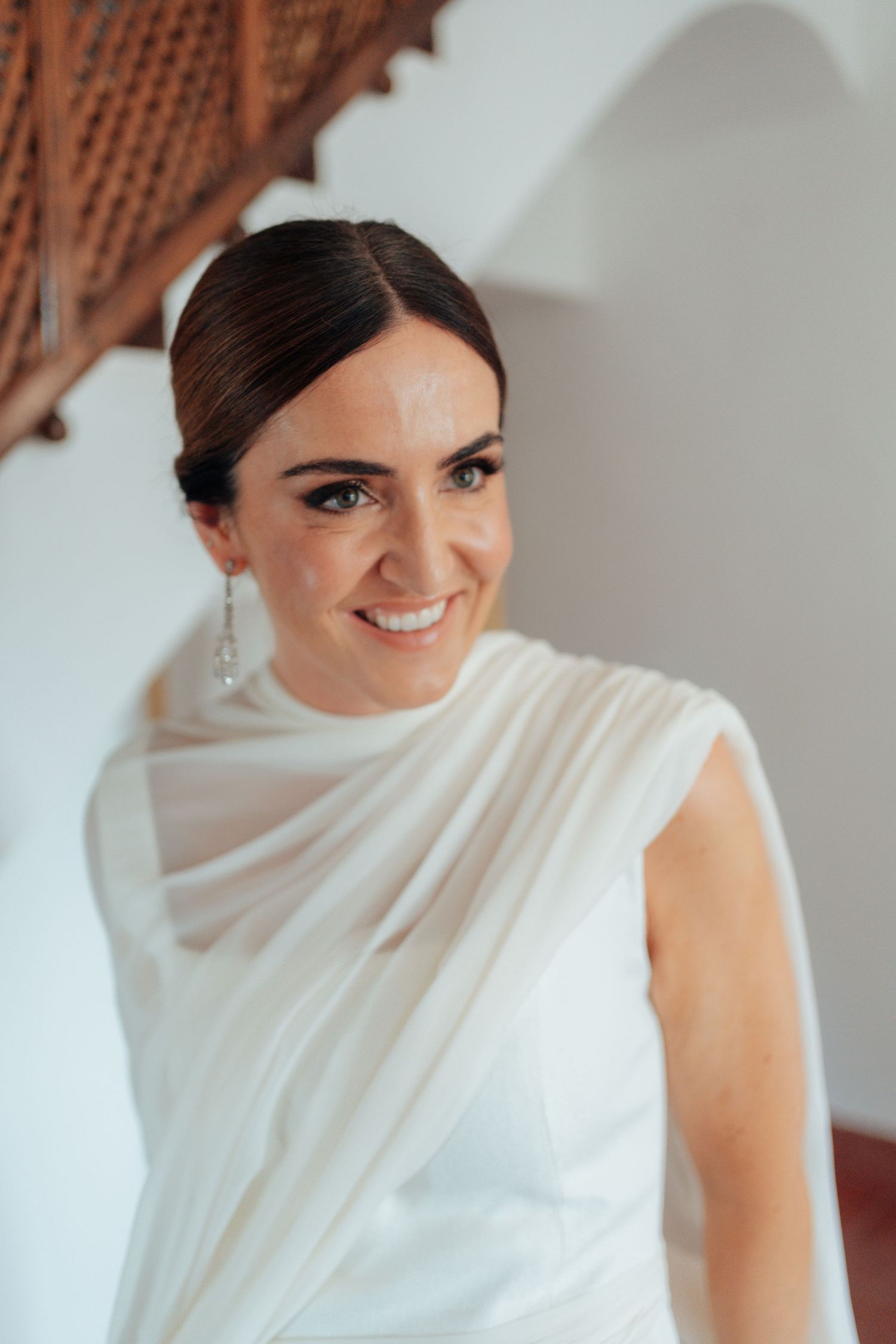Radiant bride in white draped wedding dress smiling warmly in bright elegant interior.