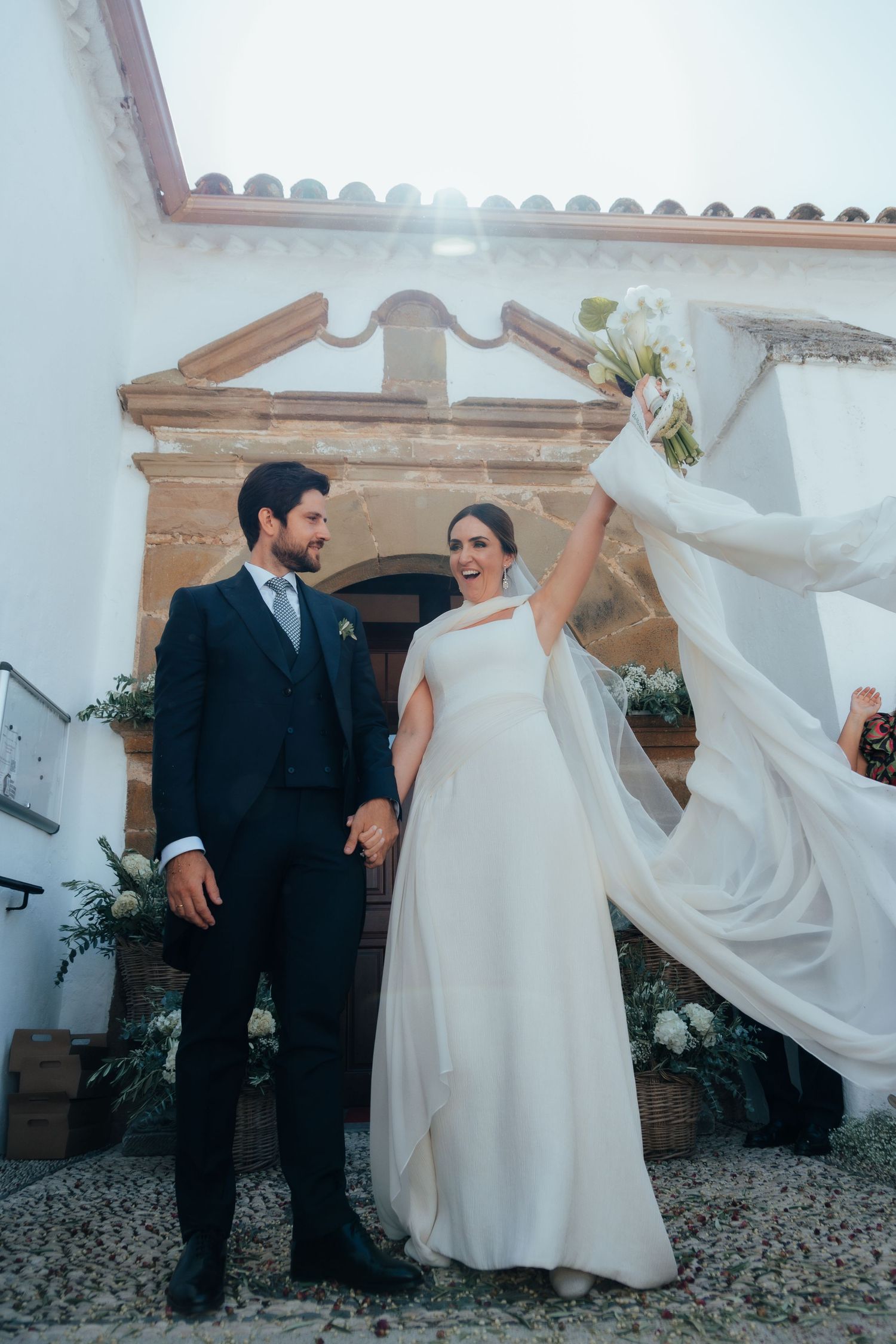 Joyful bride raising her bouquet while holding hands with groom outside a white Spanish church.
