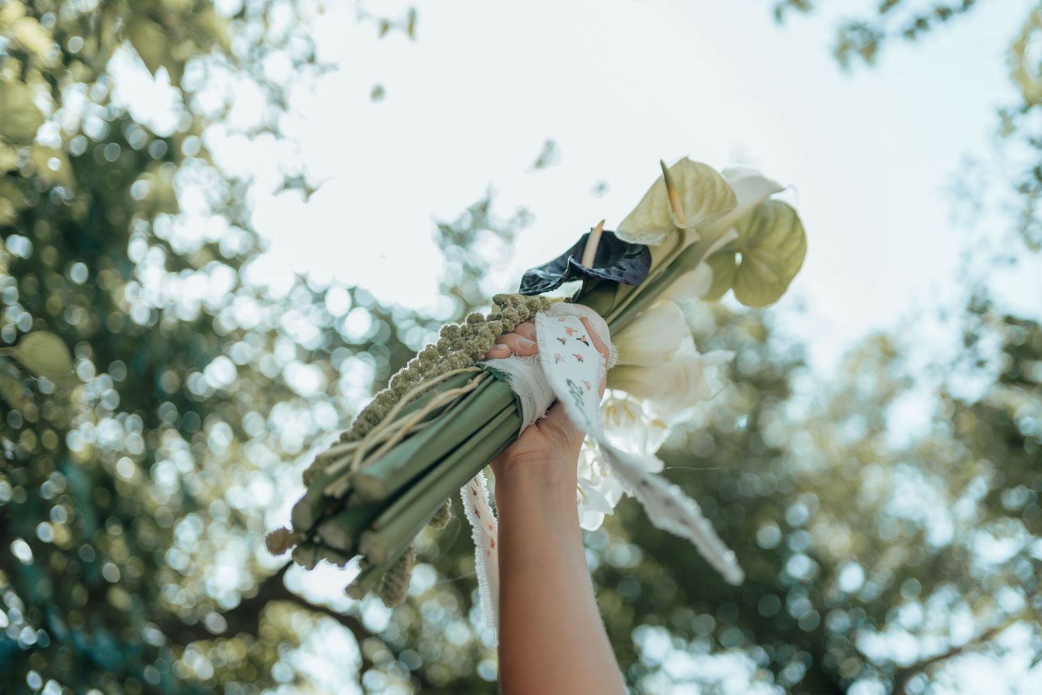 Hand raised holding a beautiful white wedding bouquet with greenery against a sunlit tree background.