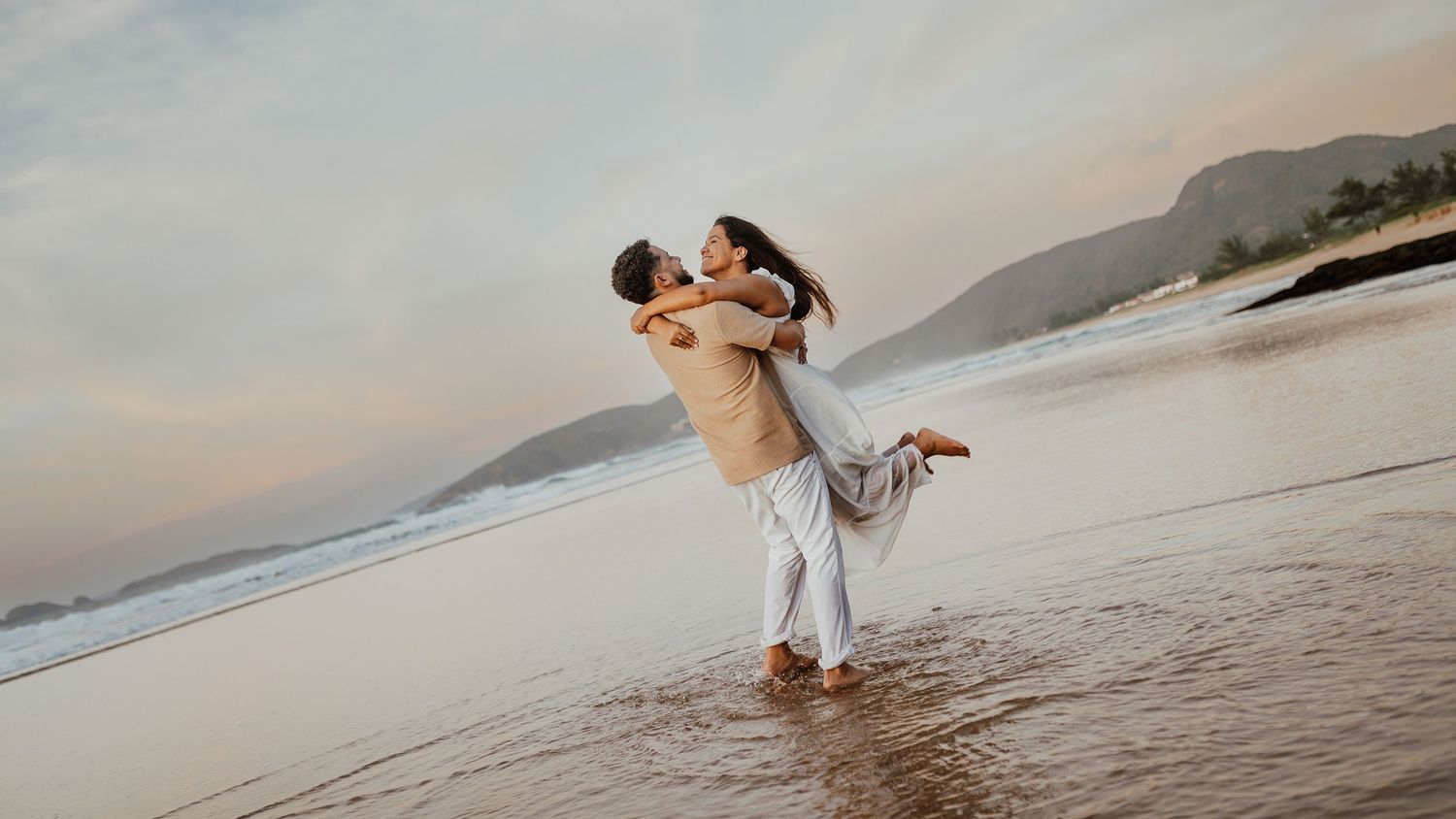 Pre-wedding romântico de Arthur e Camila na praia de Búzios ao pôr do sol. Noivo levantando a noiva na areia molhada.