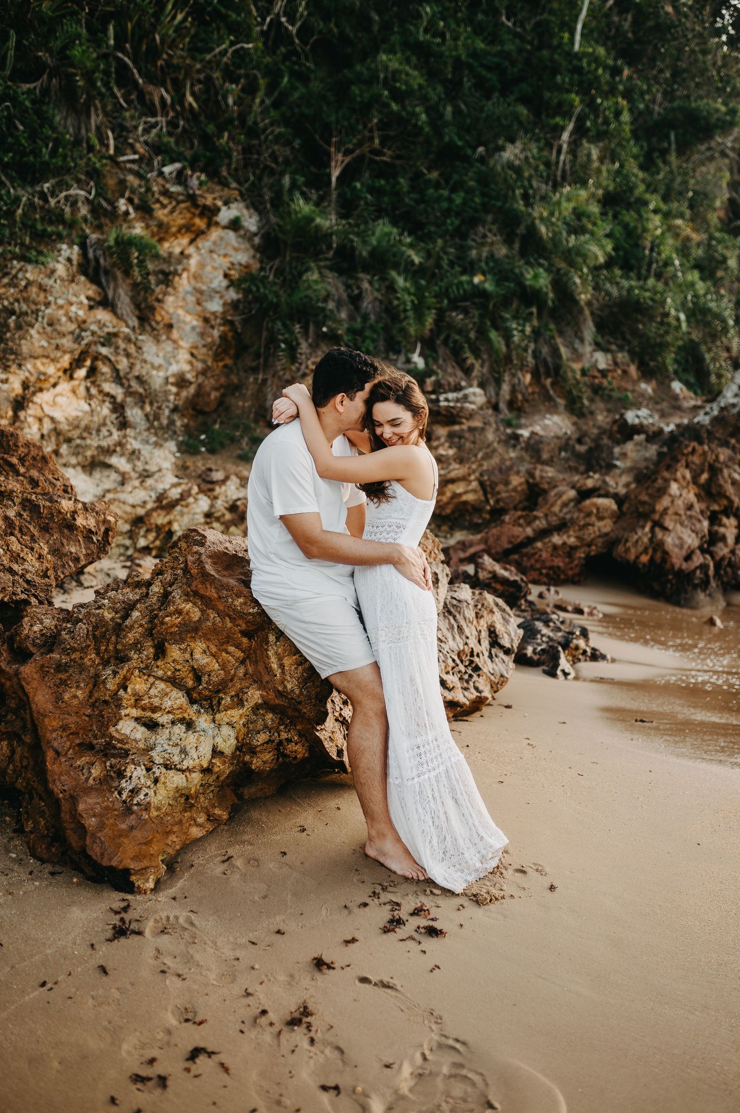 Ensaio de casal em Búzios: Bruno e Magale abraçados em pé na areia com fundo de pedras naturais na praia do Pai Vitório em Búzios - RJ