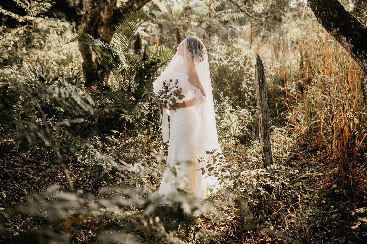 Noiva com buquê e véu longo em meio à vegetação, estilo casamento no campo, no Sitio Latitude 22 em Rio das Ostras.
