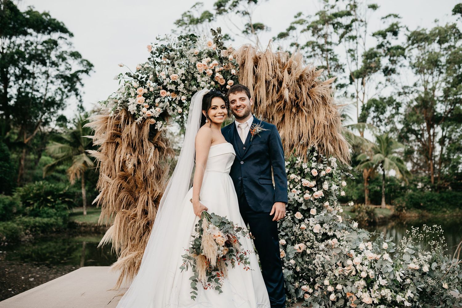 Noivos sorrindo no altar externo durante casamento na Casa do Lago em Rio das Ostras, com decoração rústica de flores e capim-dos-pampas
