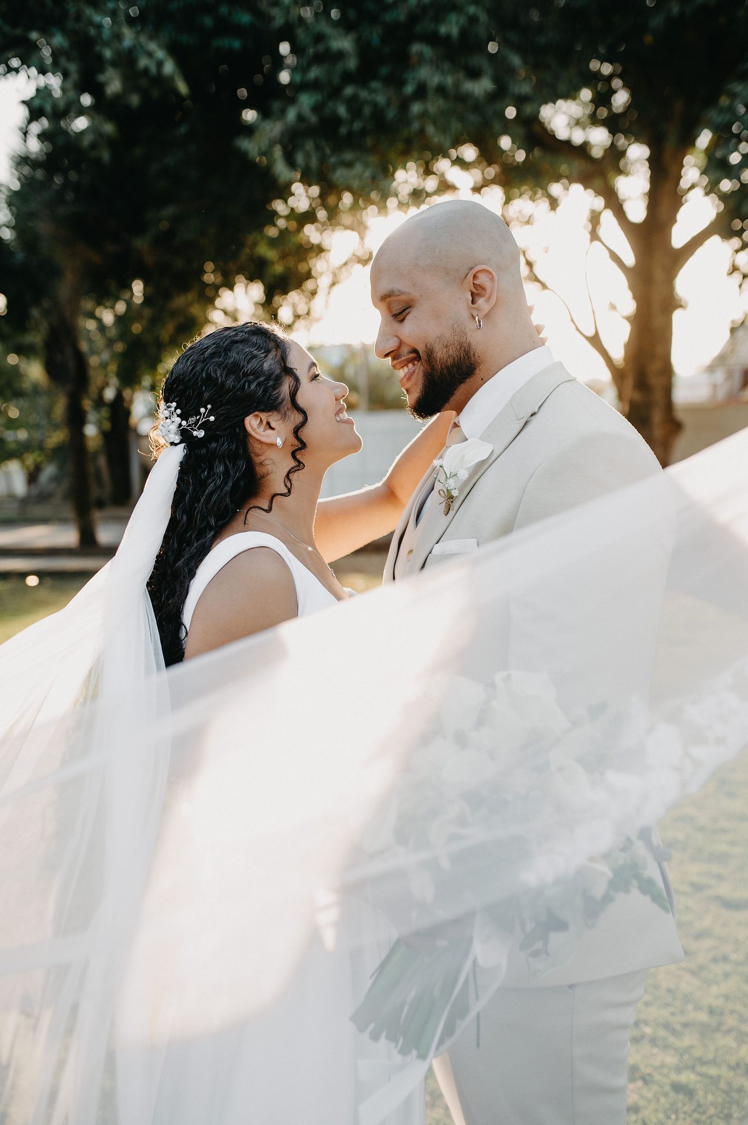 Foto de casamento ao ar livre no Sítio Boa Vista, Rio das Ostras. Noivos sorrindo em luz dourada do pôr do sol, com véu esvoaçante.