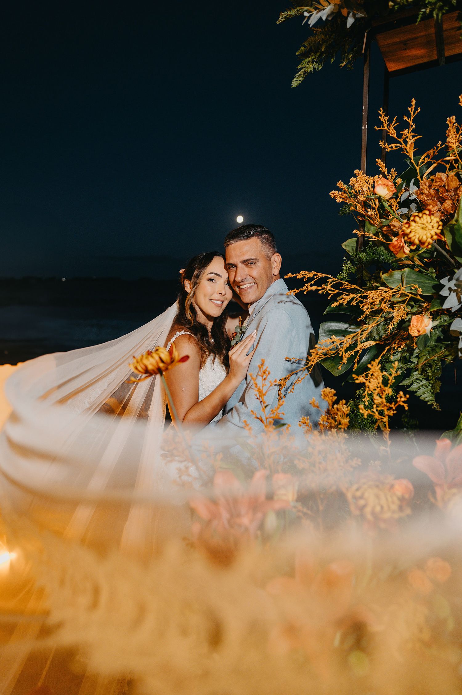 Casal de noivos abraçados em um casamento noturno no Espaço Solar em Rio das Ostras com véu esvoaçante, lua ao fundo e rica decoração floral