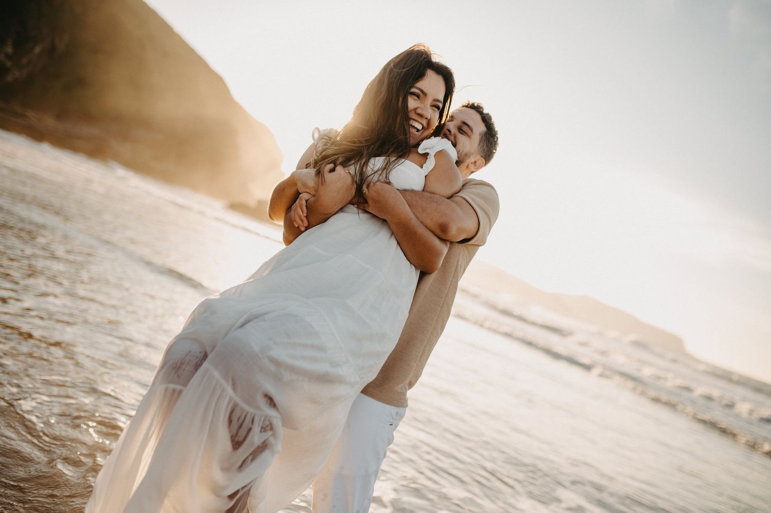 Casal abraçado sorrindo em ensaio fotográfico na praia de Tucuns em Búzios, luz natural.