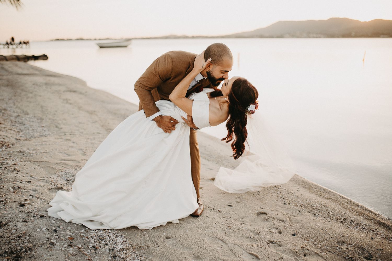 Foto romântica de casal na Lagoa de São Pedro da Aldeia ao pôr do sol. Noivo beijando a noiva com terno marrom e vestido de noiva na areia.