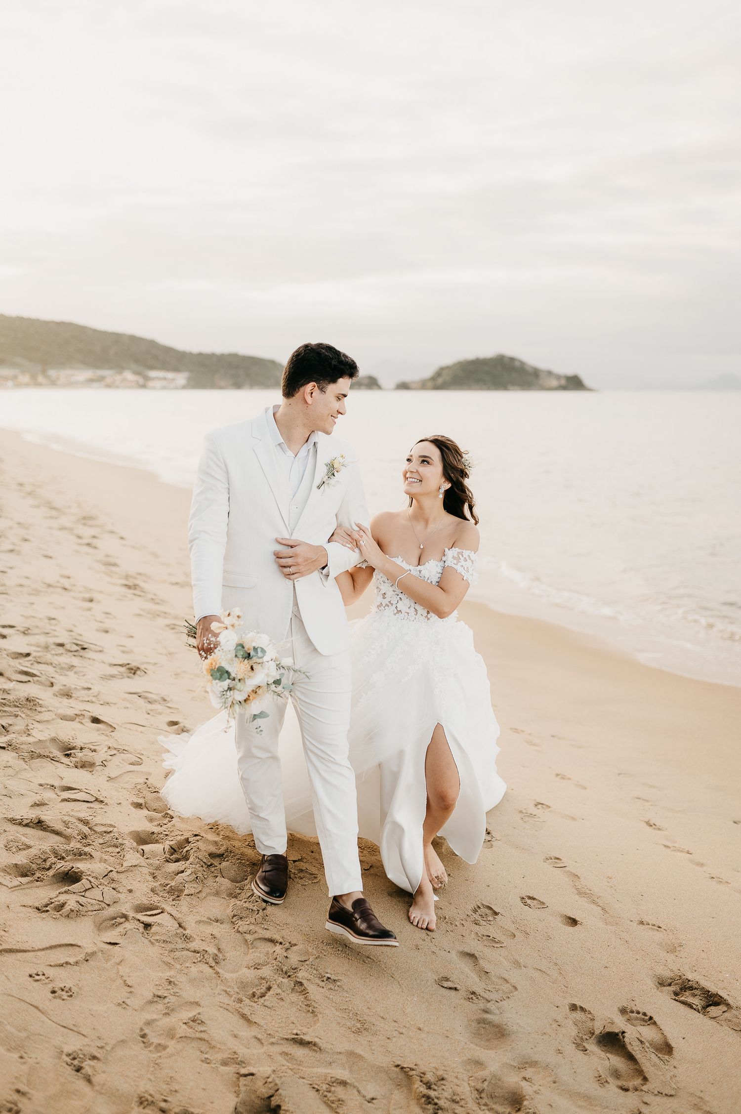 Casal de noivos recém-casados na praia deserta com o mar e morros de Búzios ao fundo, após casamento no CASAMAR.