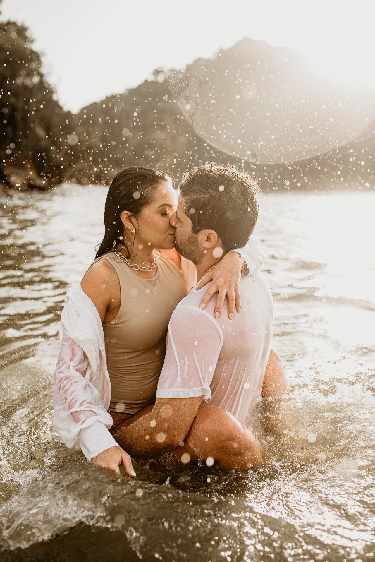 Foto artística de casal se beijando dentro do mar em Búzios. A luz do sol cria um efeito dourado nas gotas de água suspensas no ar.
