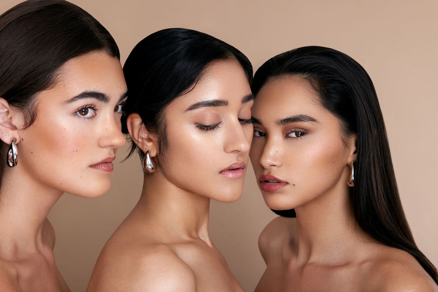 Three models with natural makeup and long dark hair pose together against a neutral beige background.