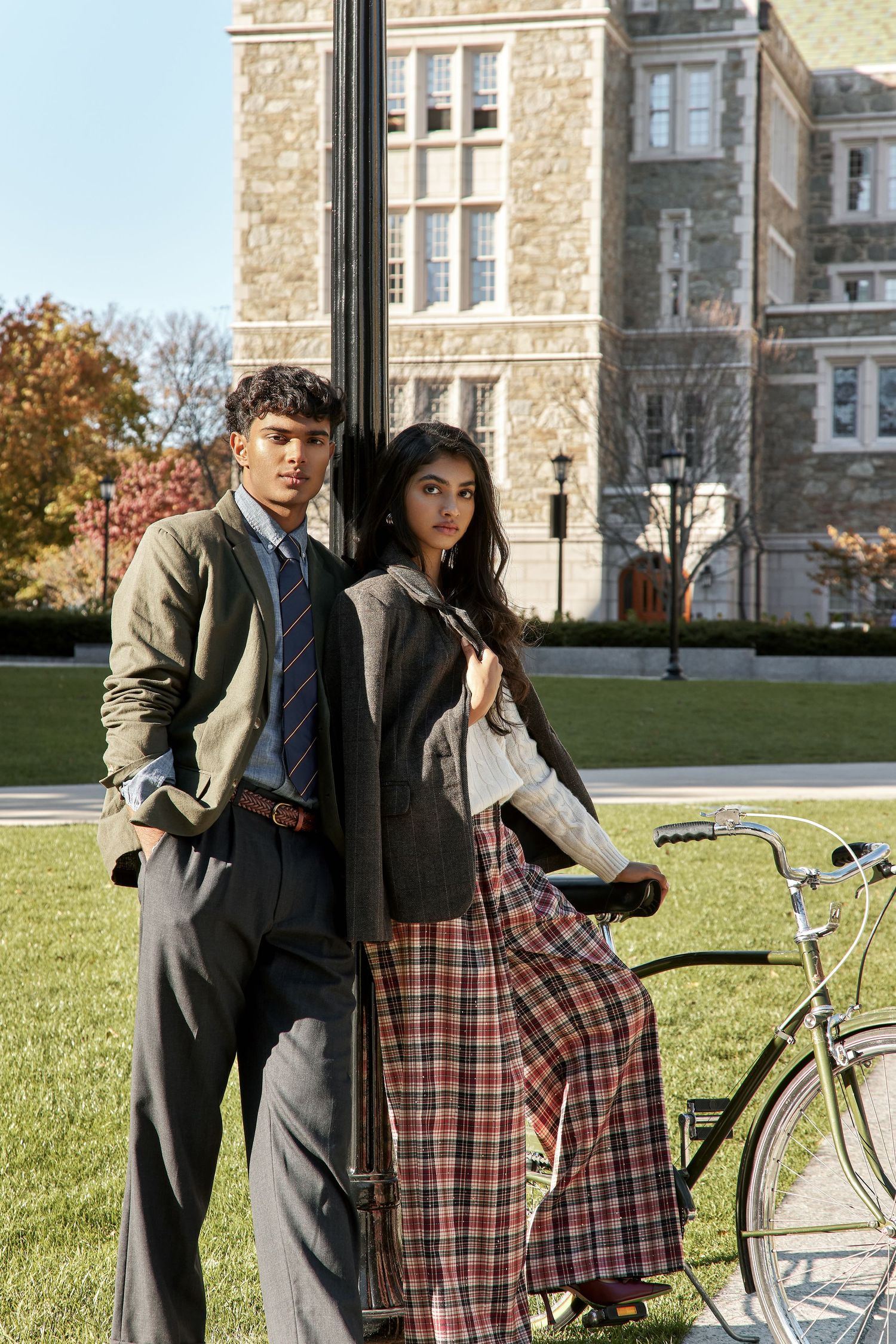 Two students in preppy collegiate attire stand on a university lawn with a vintage bicycle near a stone building.