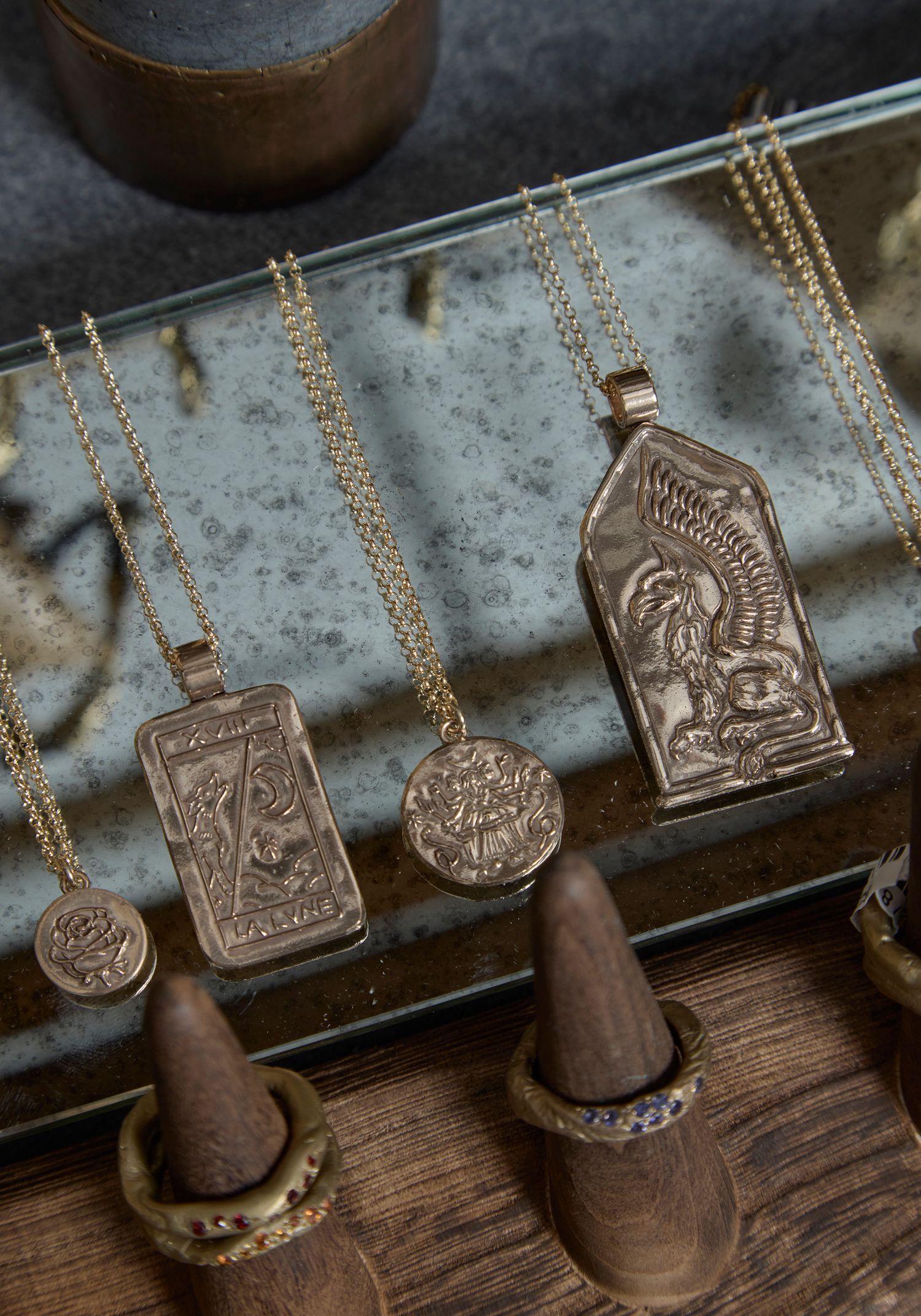 Gold pendant necklaces with antique coin and tarot card designs displayed on a rustic tray with wooden ring holders.