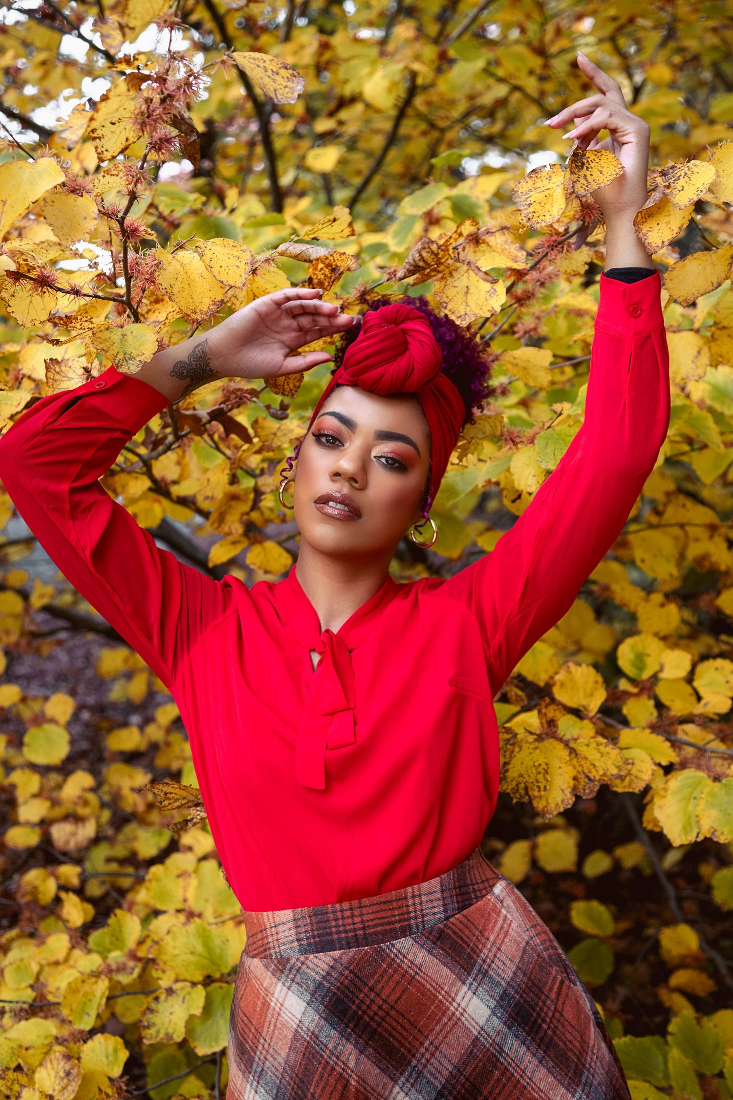 Person in bright red sweater and plaid skirt poses playfully against autumn foliage with yellow leaves in background.