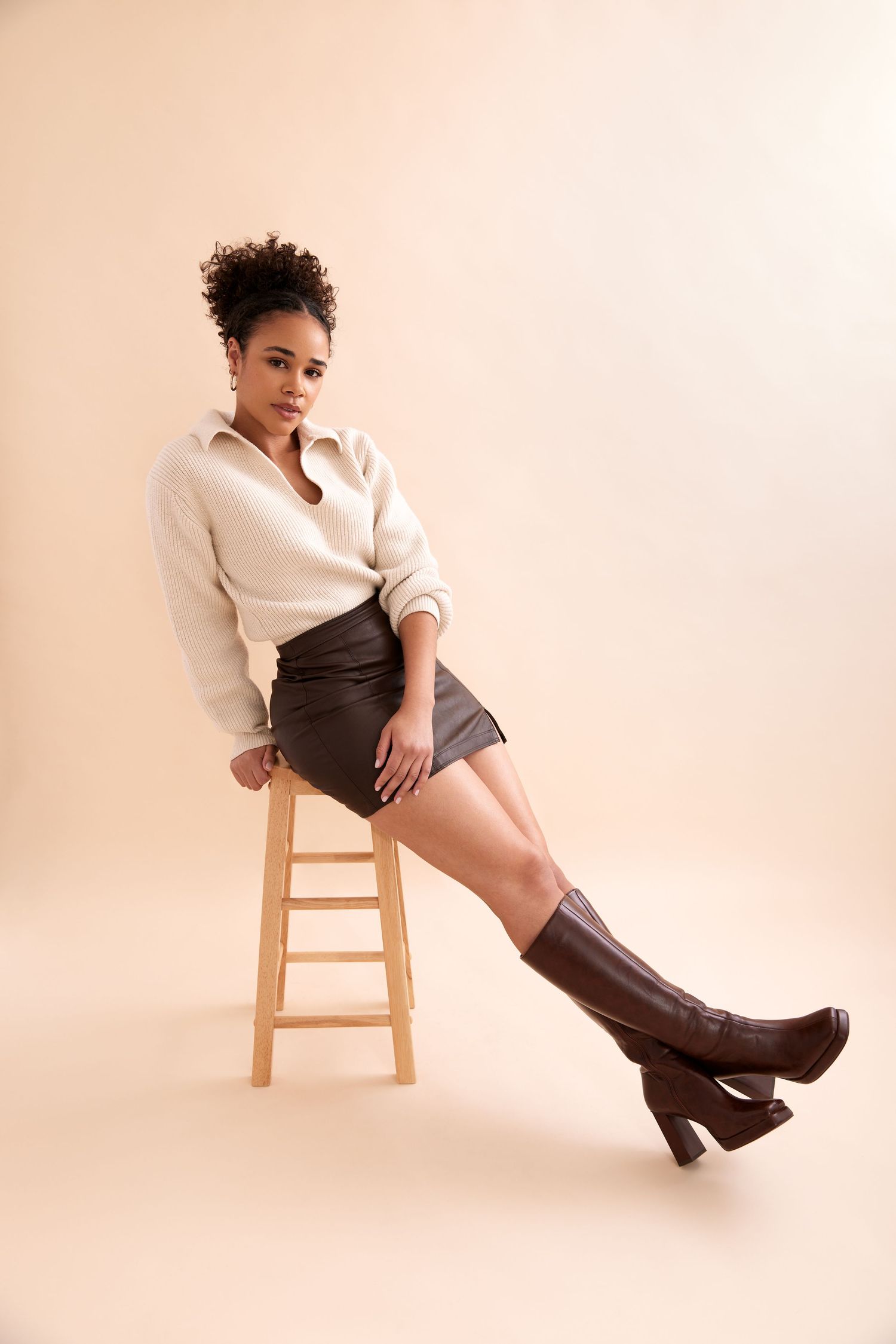 Fashion portrait with white blouse, black skirt and brown knee-high boots against beige background while sitting on wooden stool.