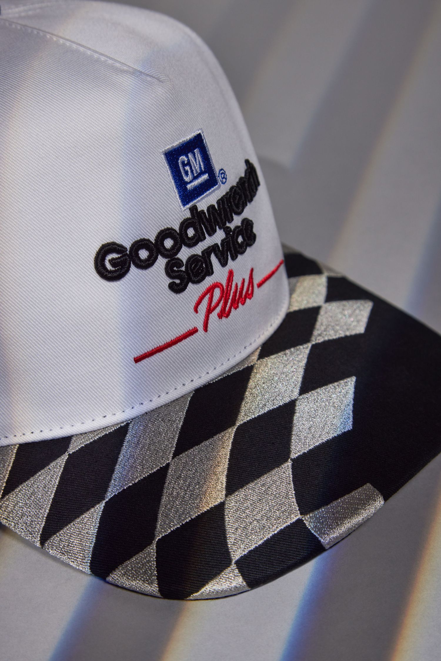 A white baseball cap with black and silver checkered pattern on the brim and Goodwill Plus logo embroidered on the front.