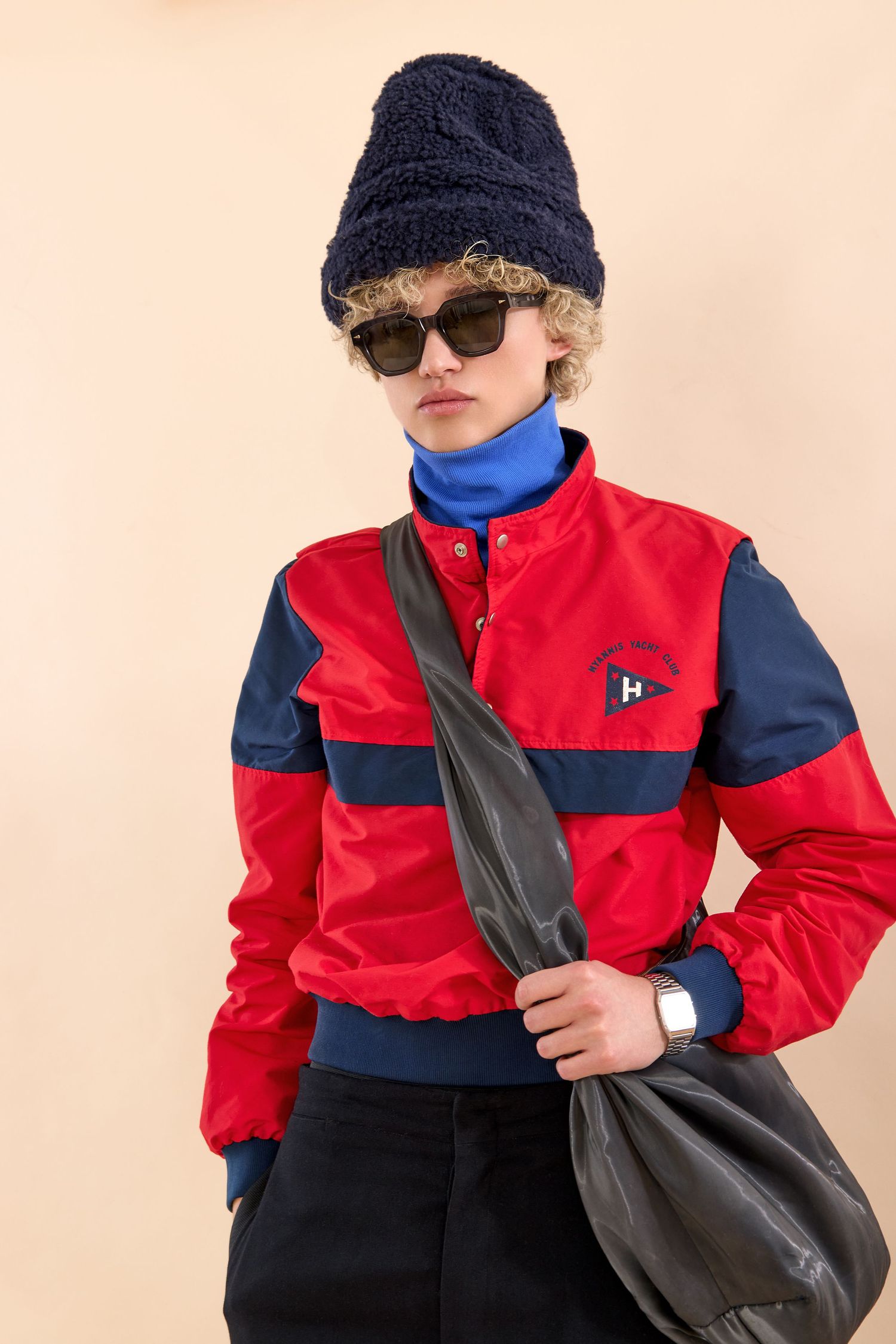 Model wearing red and navy colorblock jacket, blue turtleneck, black beanie, sunglasses, and leather bag.