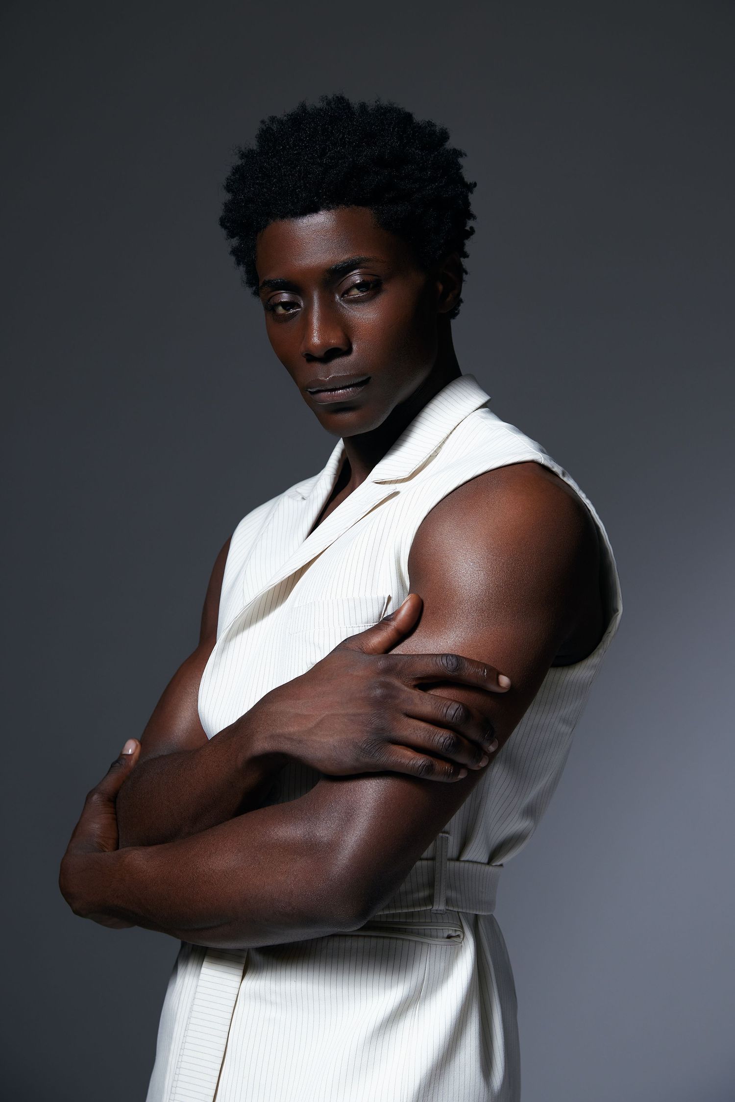 Studio portrait against dark background showing person in sleeveless white outfit with arms crossed in confident pose.