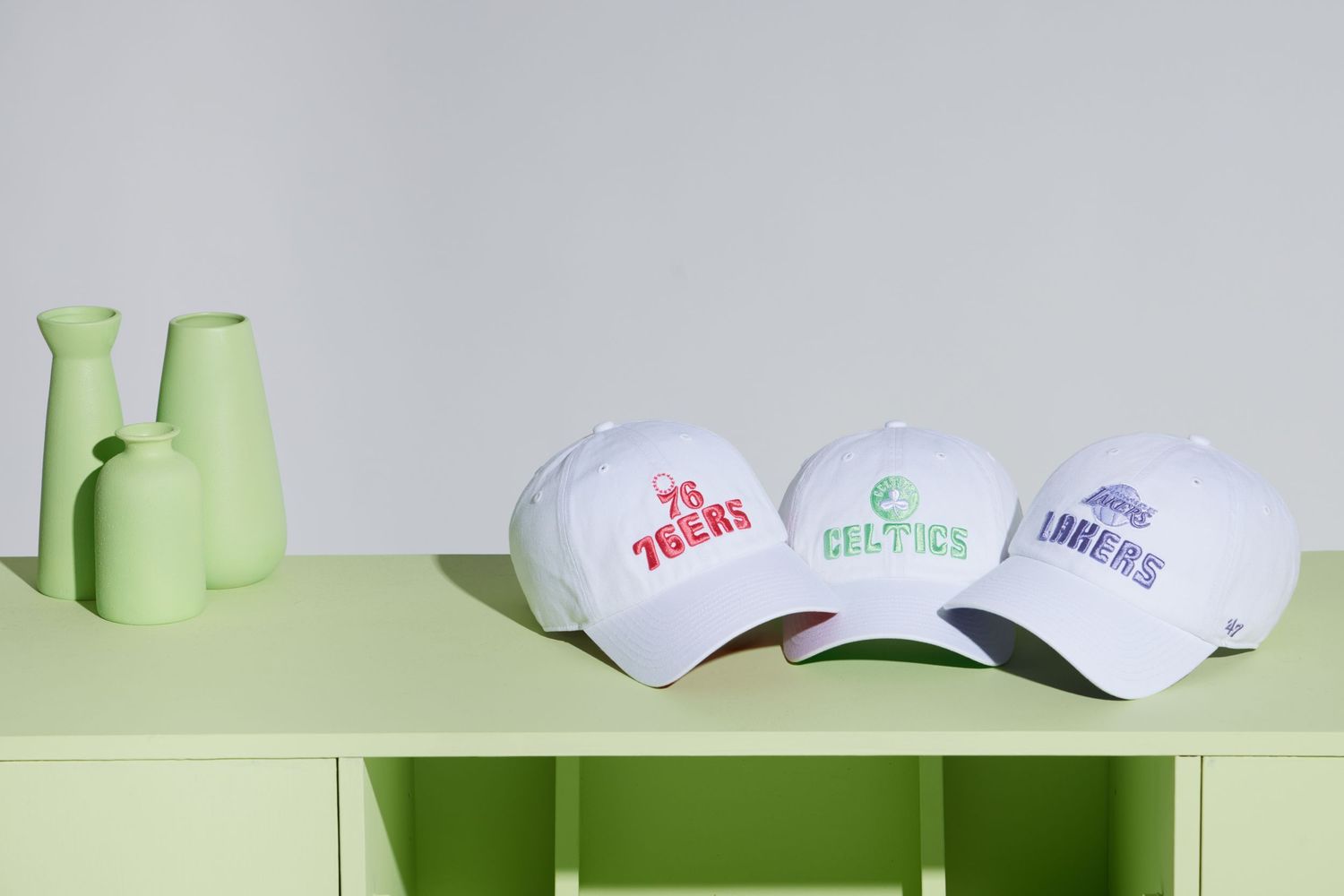 Three white baseball caps with different logos displayed on a green surface next to light green ceramic vases.