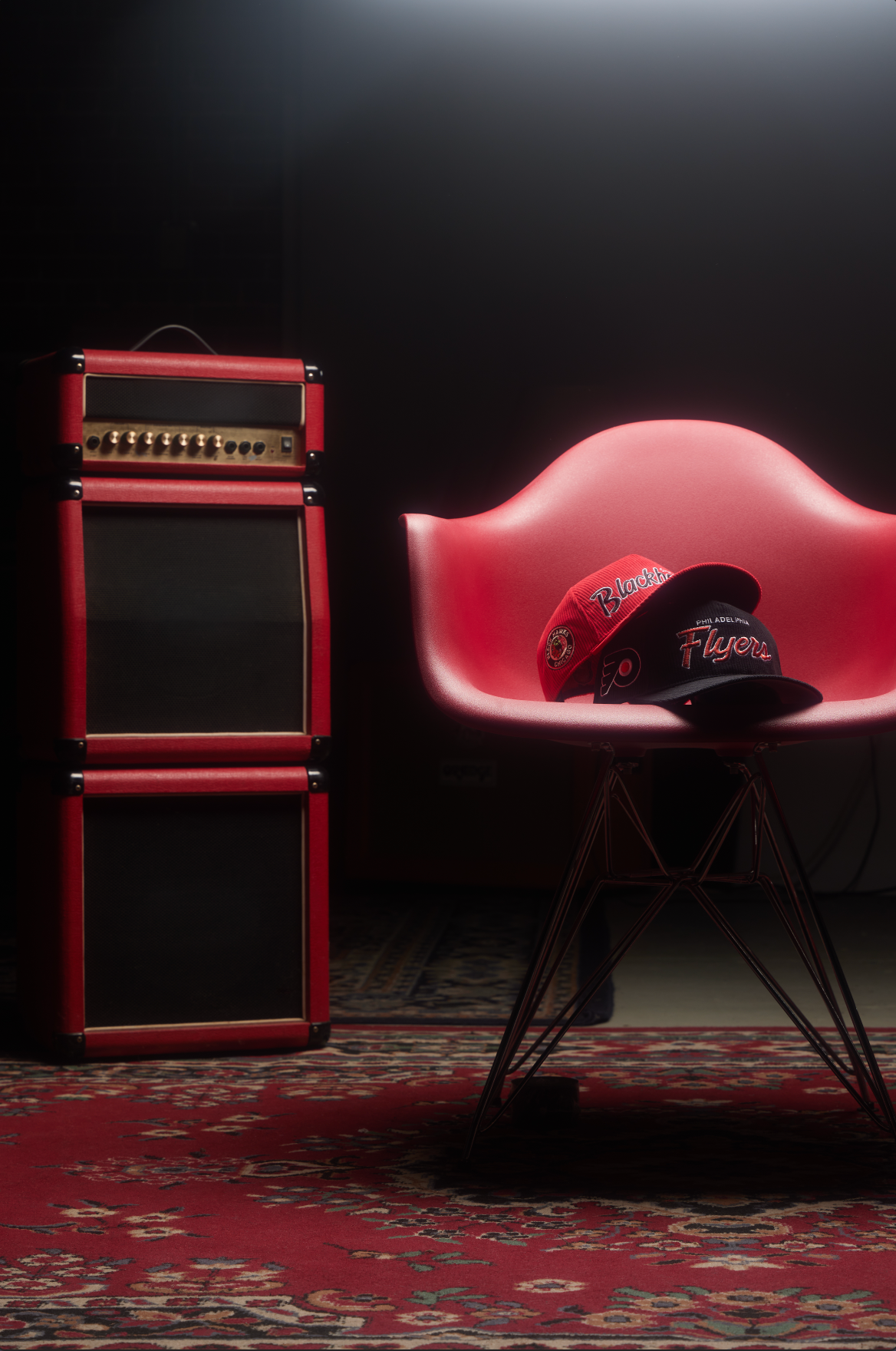 Red guitar amplifier stack and modern chair on patterned carpet in dark moody lighting.