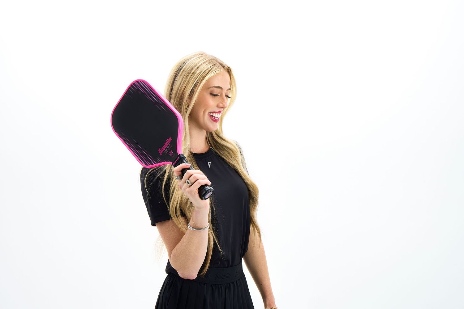 Woman in black dress and pink heels talks on phone while walking with a pickleball paddle