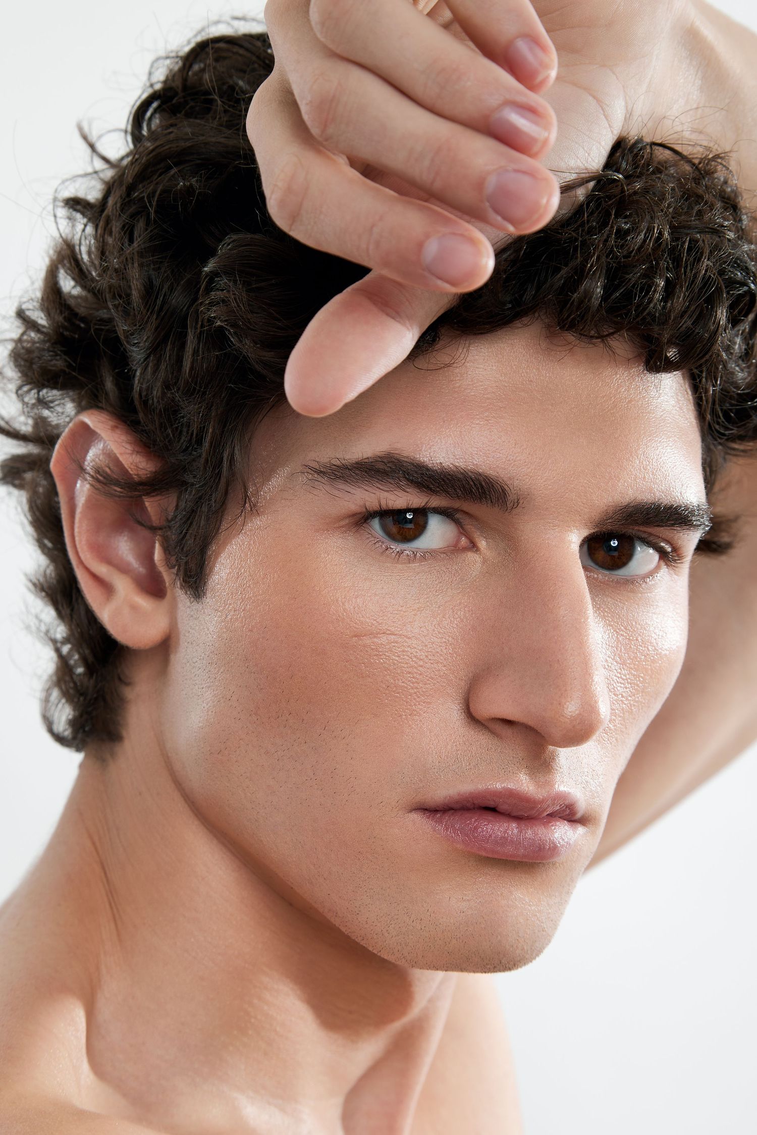 Close-up beauty shot showing curly dark hair and defined facial features in a minimalist studio setting.