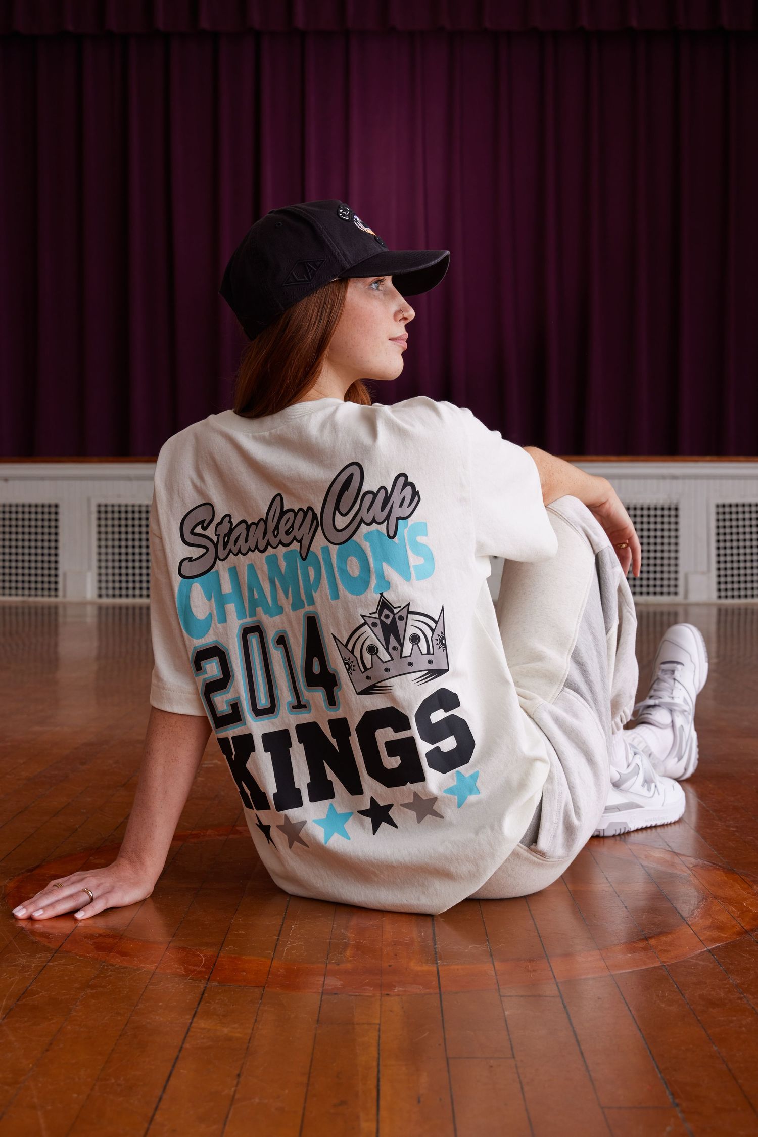 Person wearing a 2014 Stanley Cup Champions LA Kings t-shirt sits on wooden floor against red curtain backdrop.