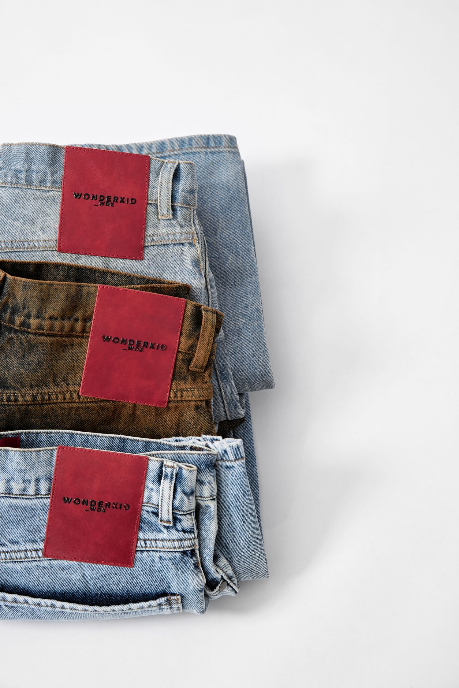 Three folded jeans with red Wonderkid labels stacked on a white surface, featuring light and dark denim styles.