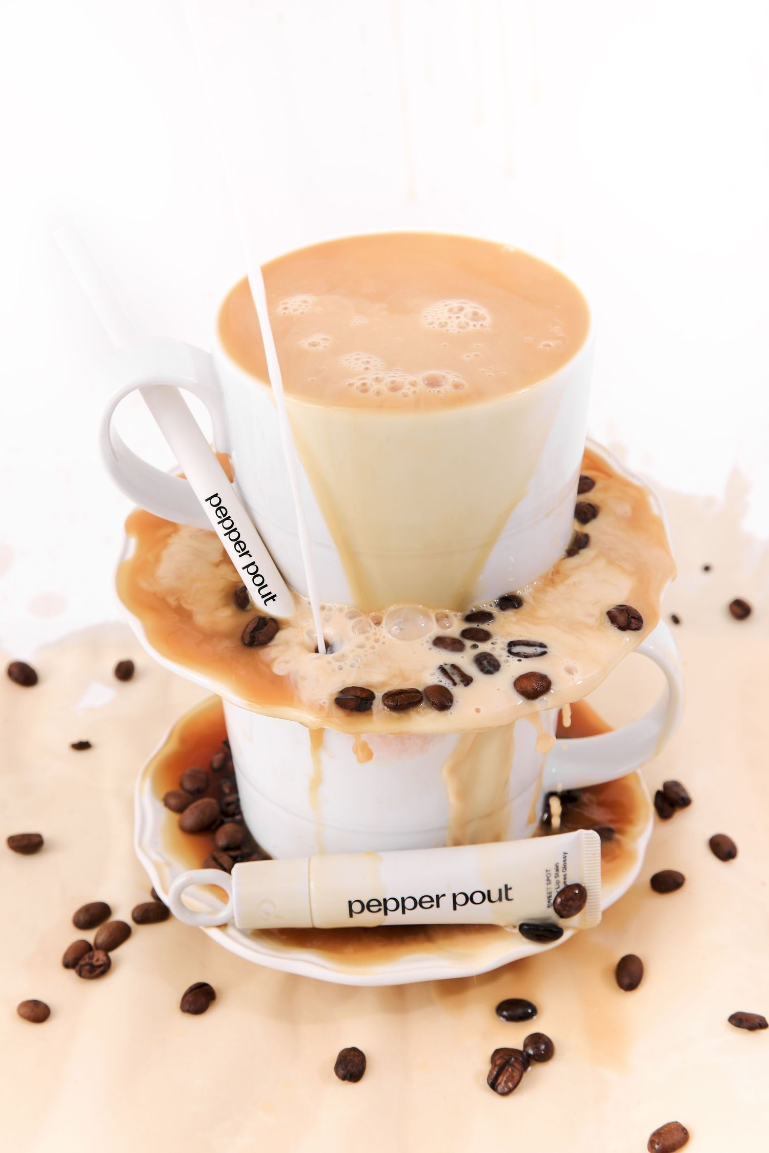 Two stacked coffee cups with spilled latte and Pepper Pout lip balm sticks surrounded by scattered coffee beans.