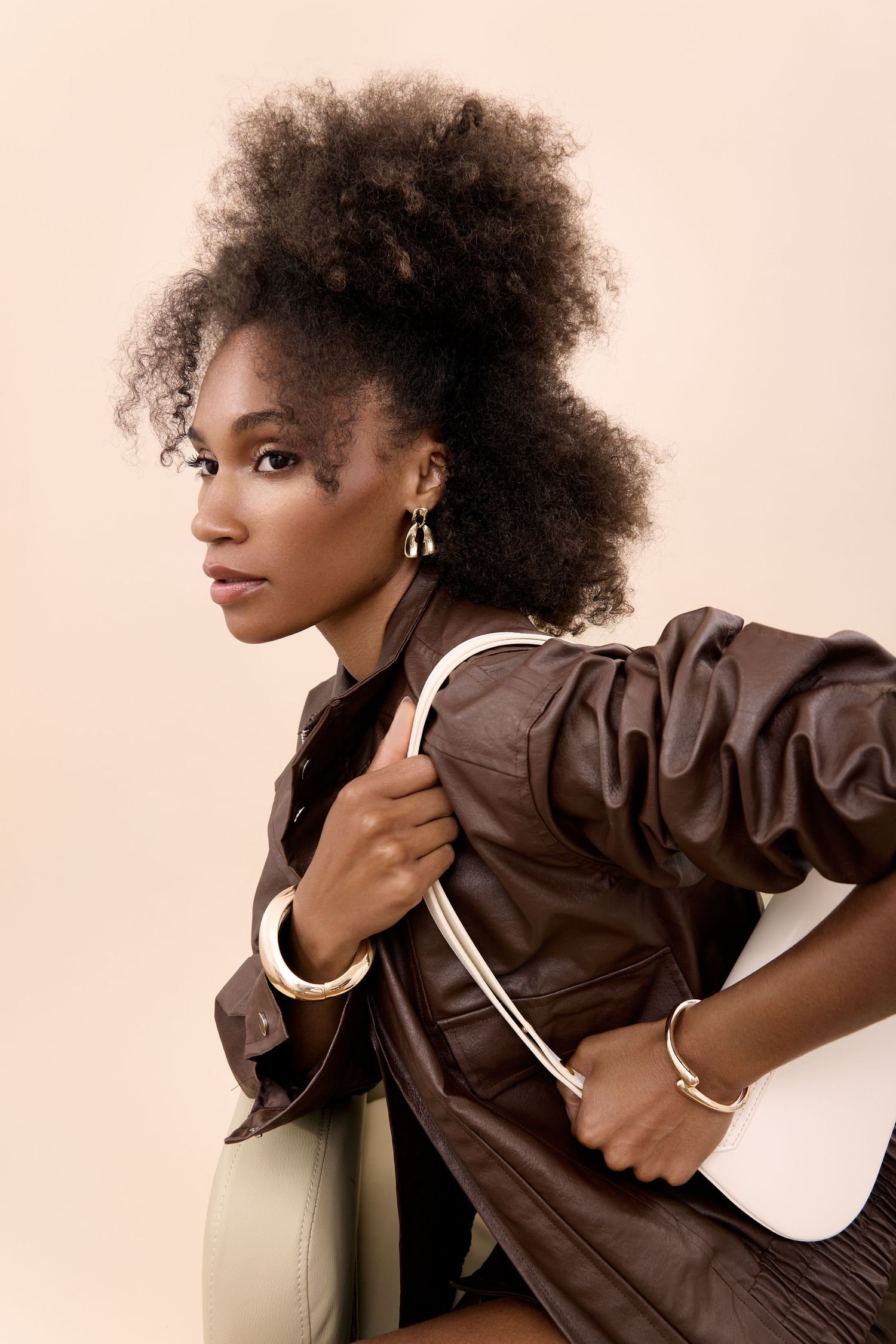 A stylish Black woman with an afro updo wears a brown leather jacket and holds a white bag in a studio setting.