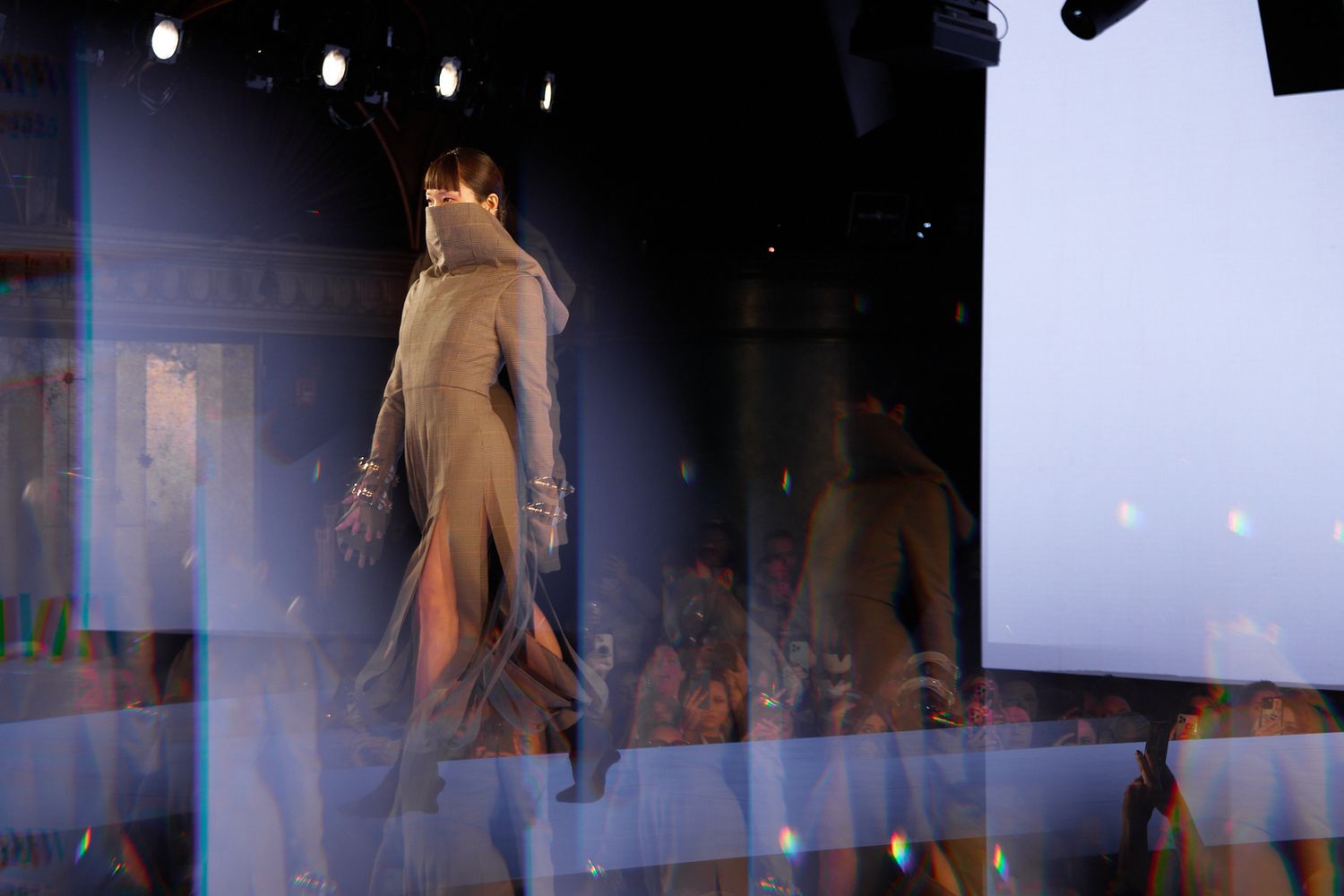 Models walk down a runway during a fashion show with dramatic lighting and smoke effects in the background.
