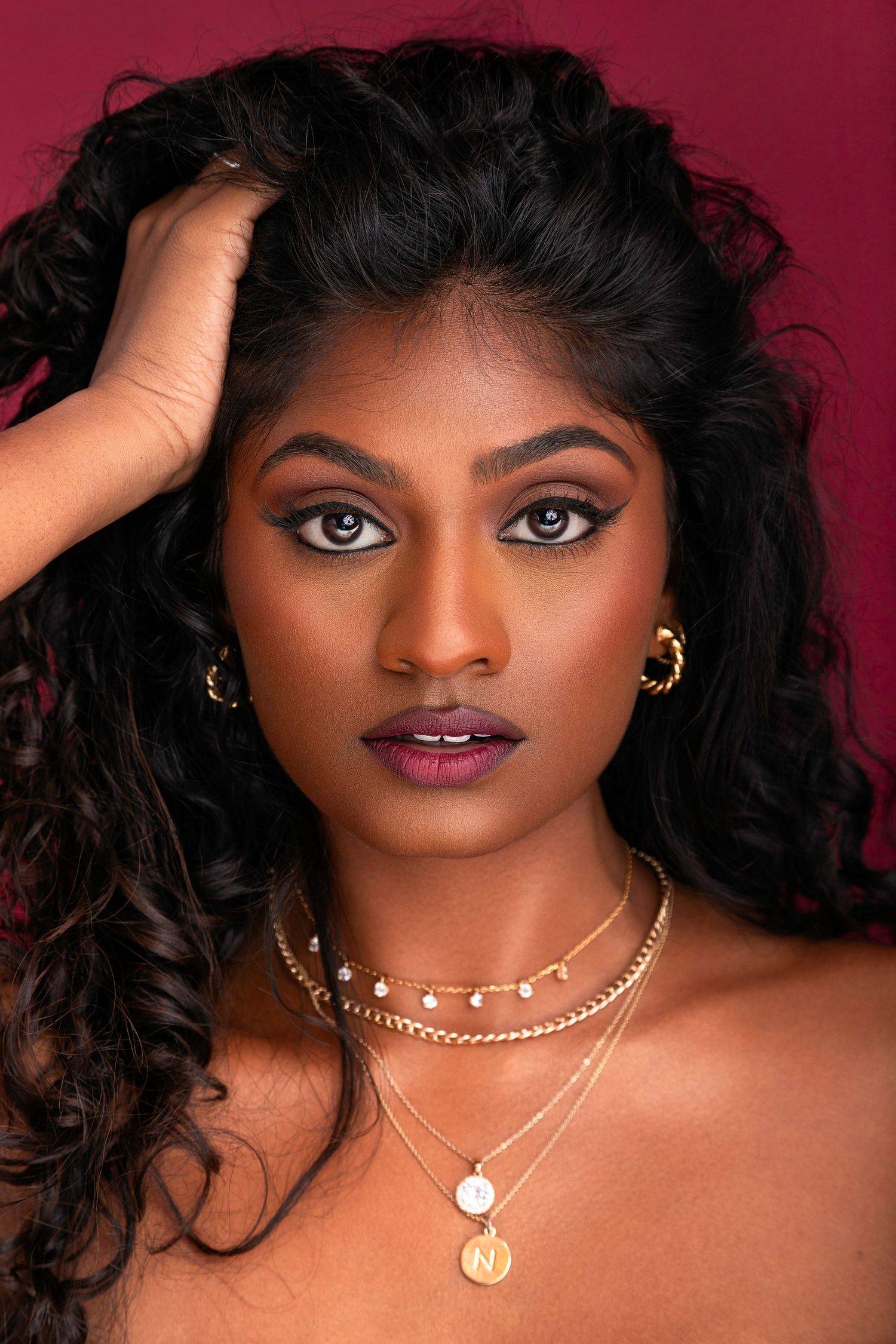 A glamorous beauty portrait with gold jewelry and dark curly hair against a deep burgundy background.