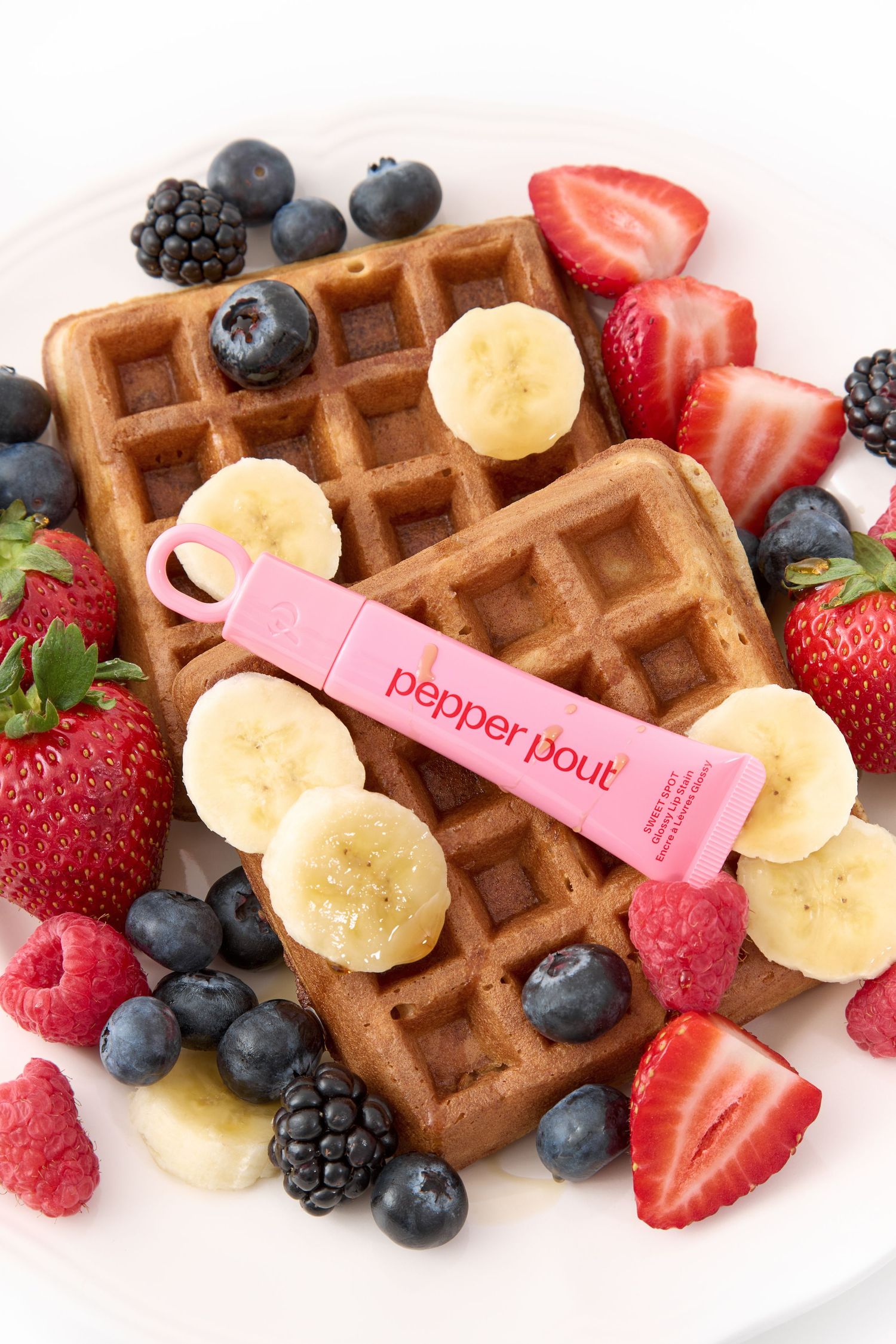 Pepper Pout lip gloss resting on golden waffles surrounded by fresh strawberries, blueberries, raspberries, blackberries, and banana slices.
