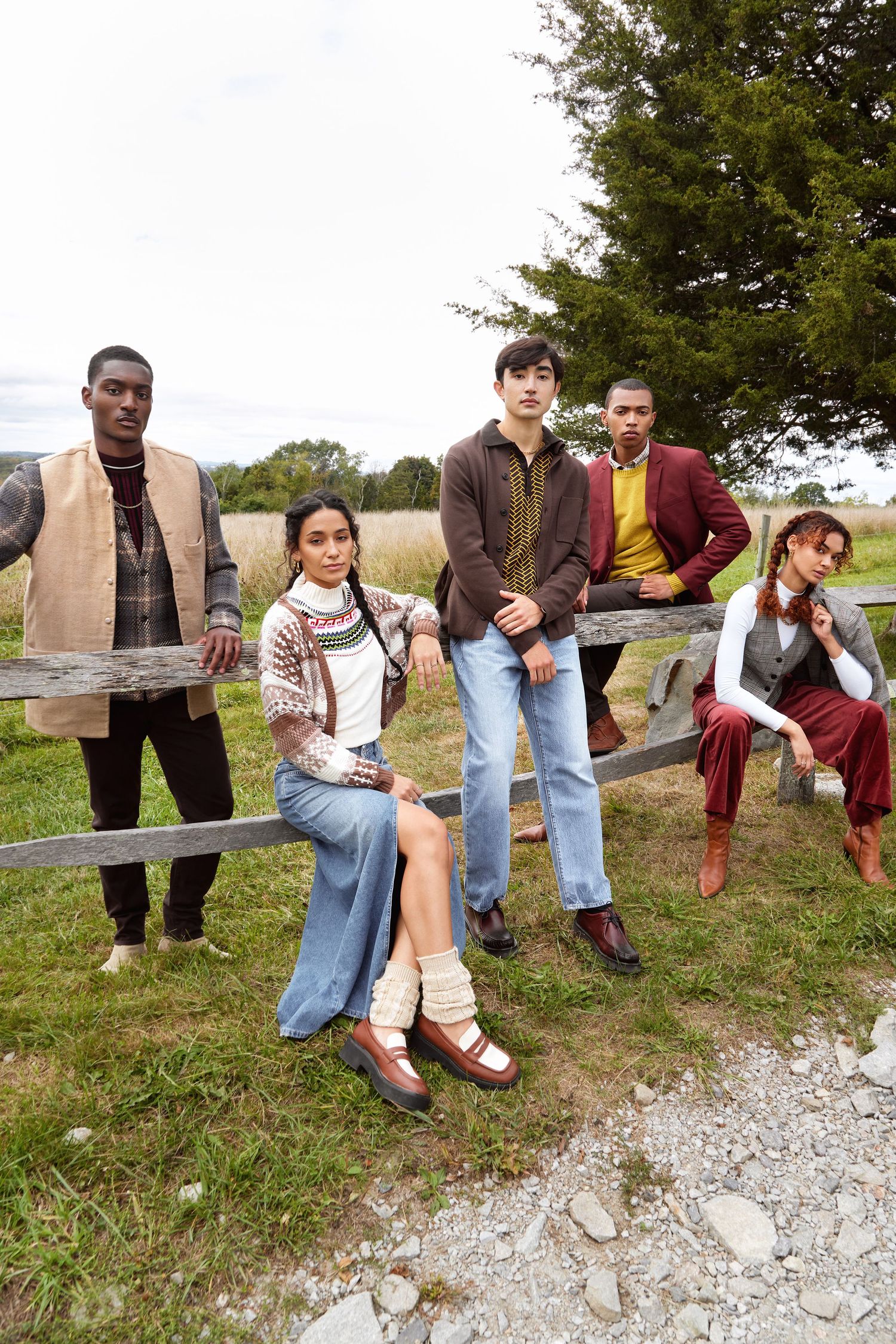A group of friends pose playfully while sitting on a gravel path near a grassy field on a sunny day.