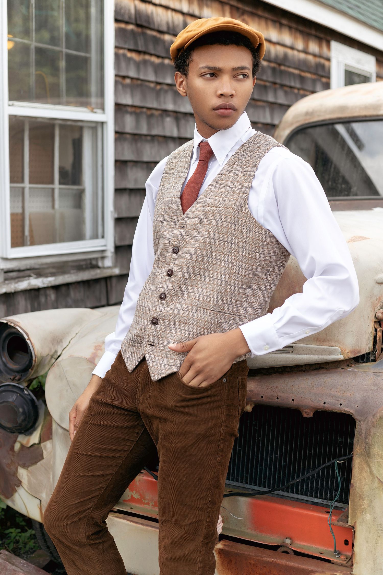 Vintage-inspired outfit featuring brown trousers, plaid vest, white shirt and cap against rustic backdrop with classic car.