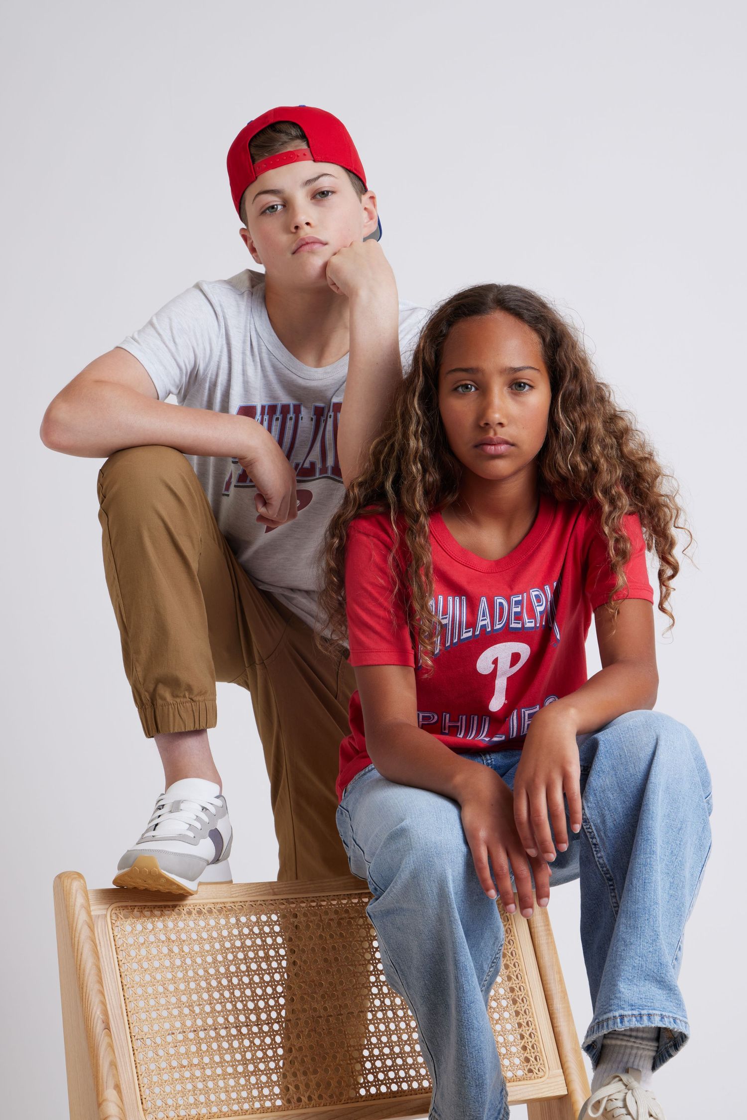 Two young people in casual clothing sitting on a woven chair against a white background.