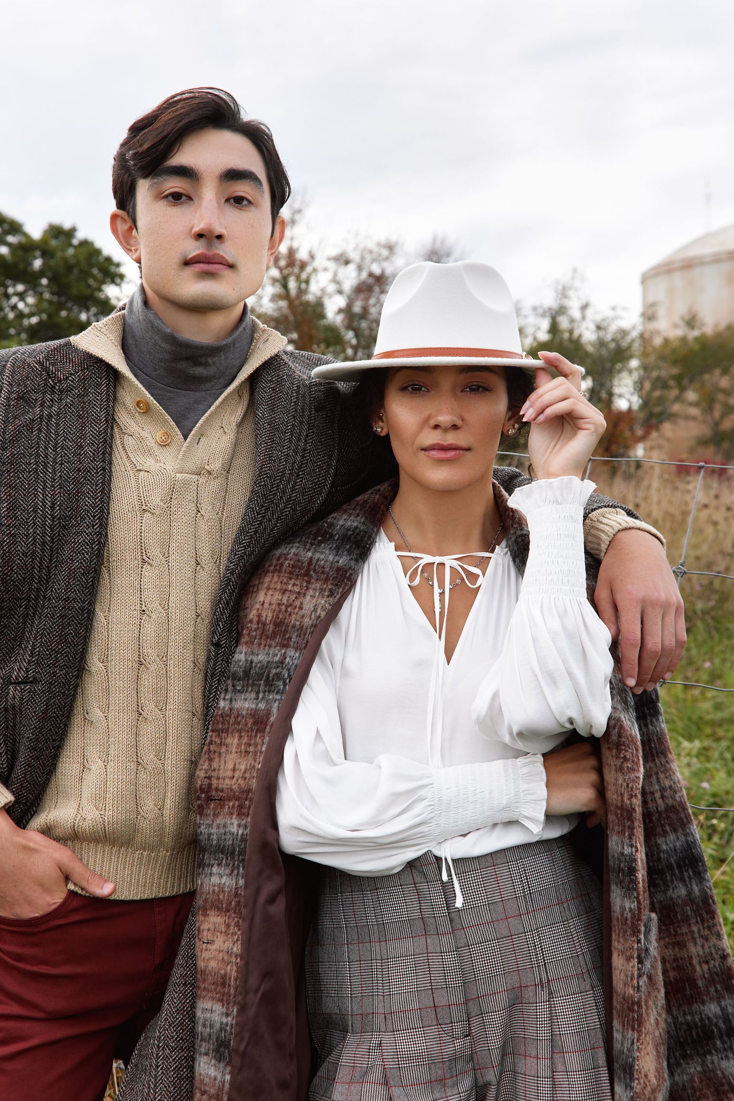 Two people in autumn fashion wearing sweaters, plaid coat and fedora hat pose together outdoors.